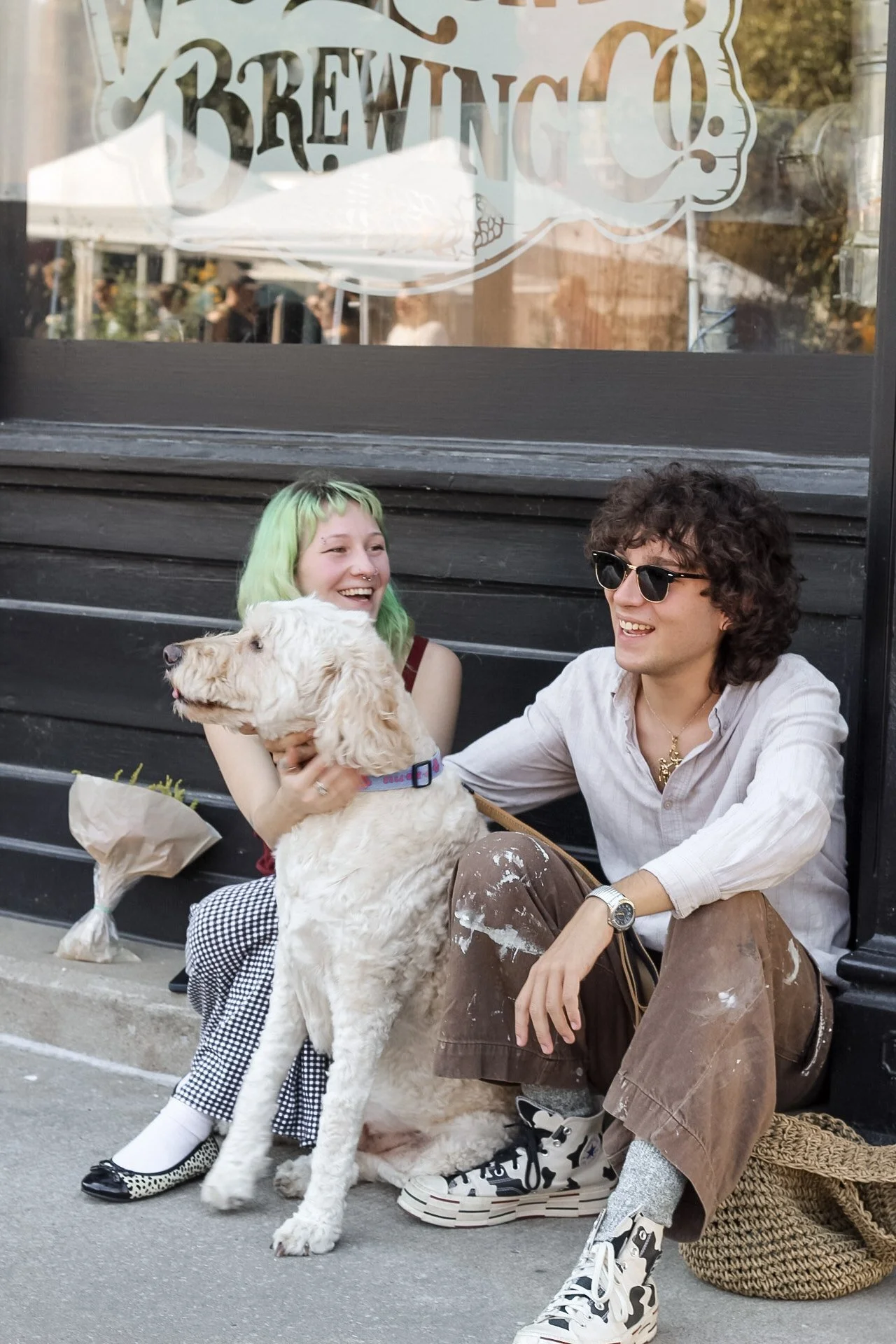 Two young adults sitting on a bench outside, smiling and laughing, with a large white dog. The woman with green hair is holding the dog, and the man wearing sunglasses is touching the dog. They are in an outdoor setting with people and trees in the b
