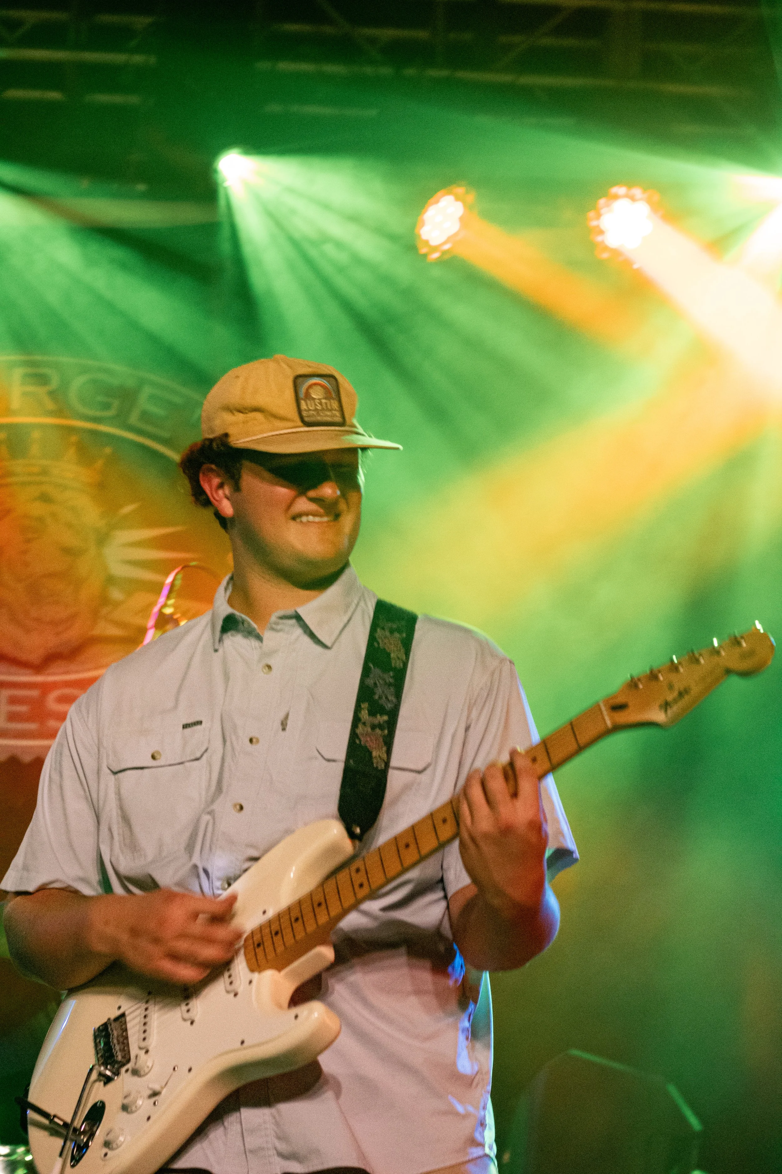 A young man with curly hair wearing sunglasses, a beige cap, and a light-colored shirt, playing a white electric guitar on stage with colorful green and yellow lights in the background.