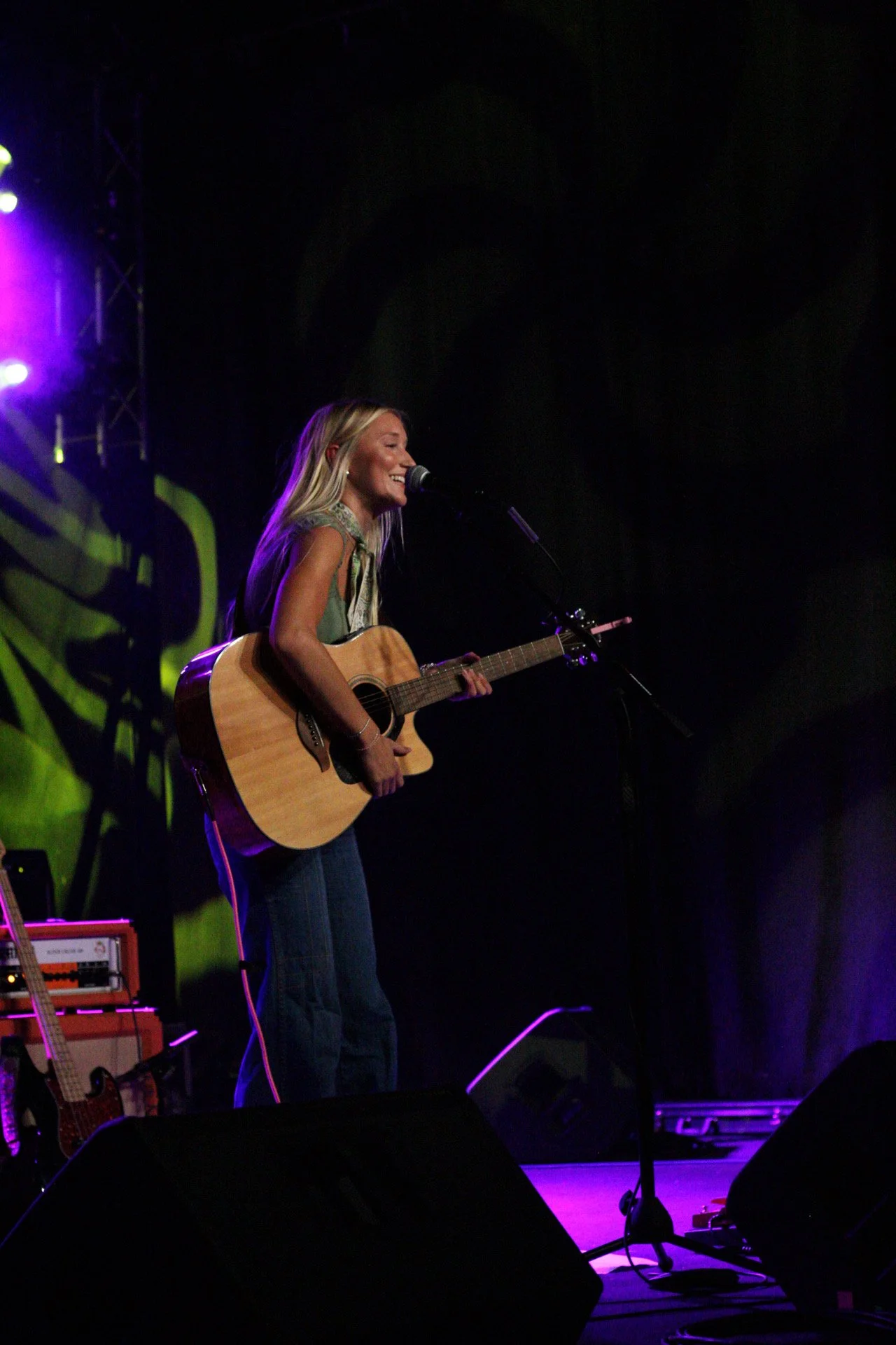 A woman performing on stage with a guitar, singing into a microphone, with stage lights and equipment visible in the background.