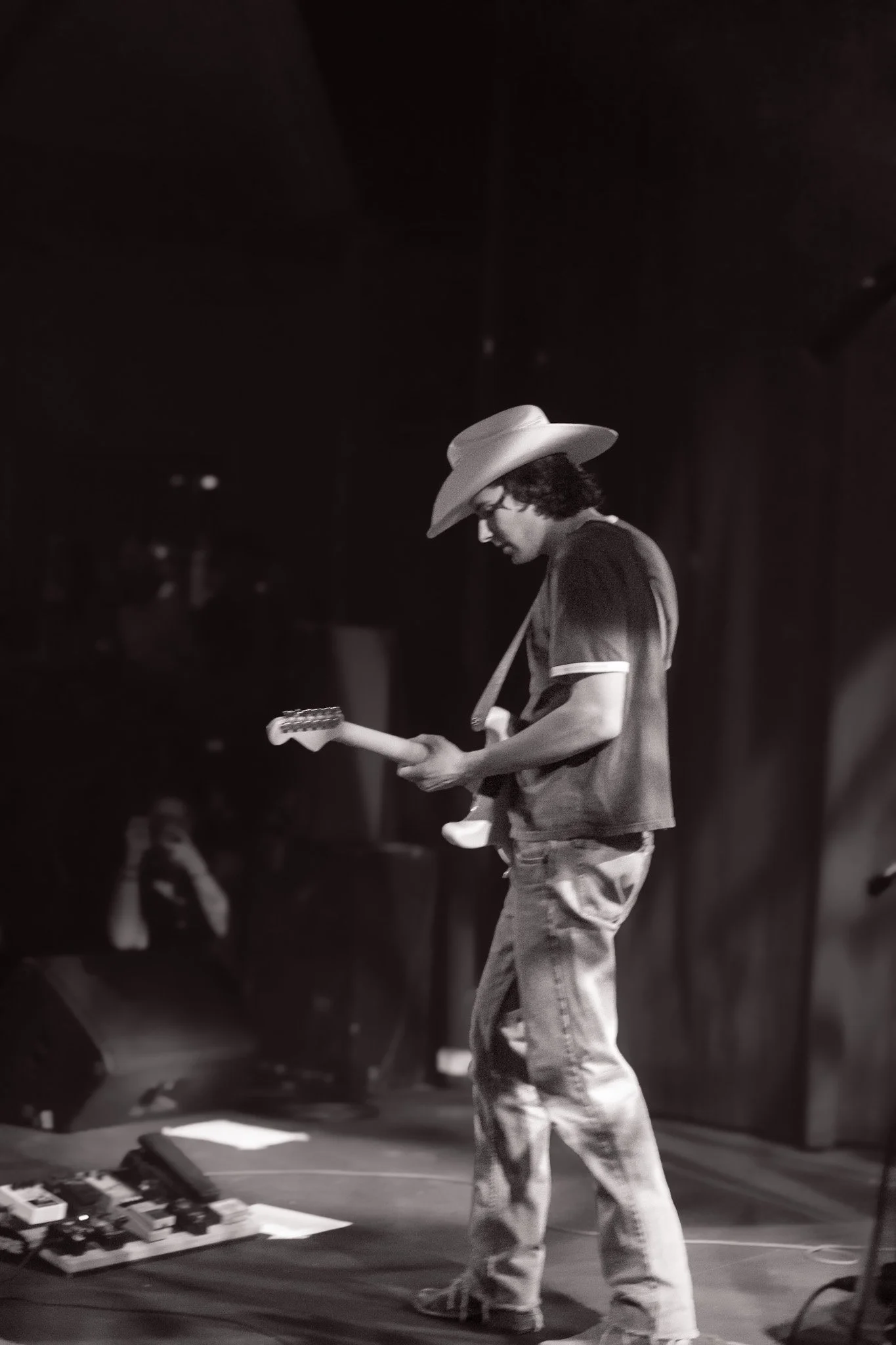 A young man wearing a cowboy hat, glasses, jeans, and a t-shirt playing an electric guitar on stage, with an audience in the background.