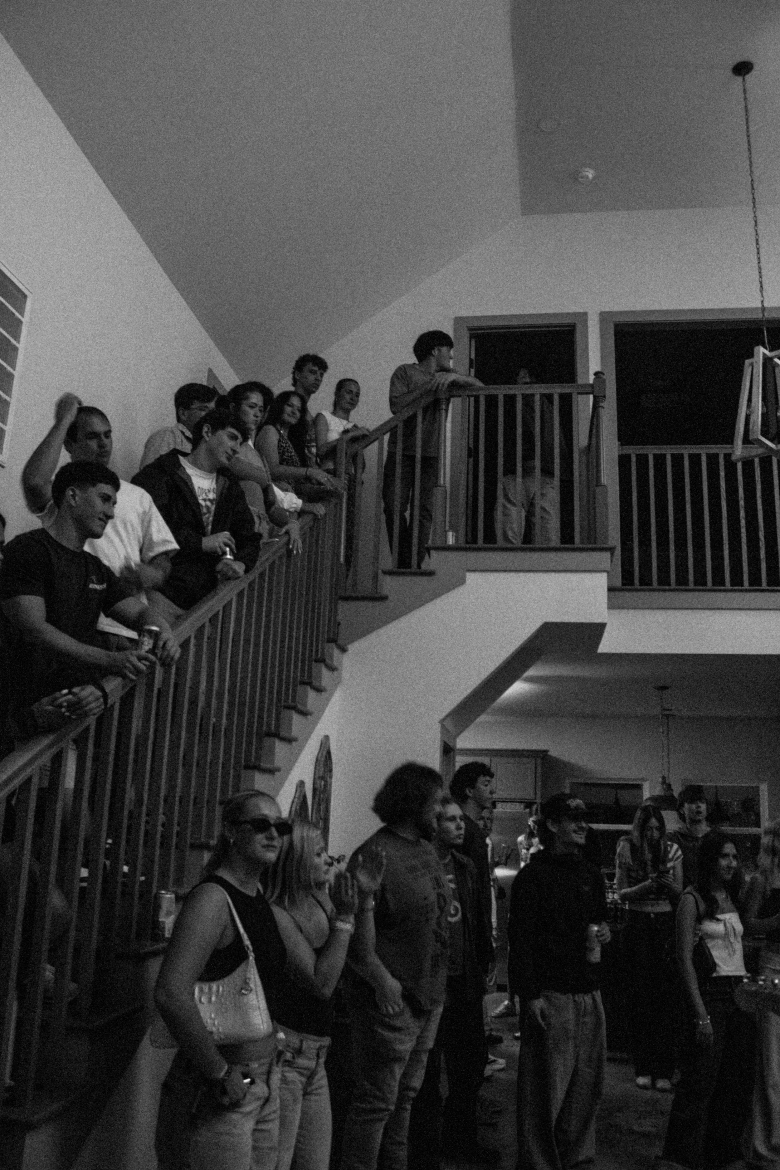 People gathered on a terrace at night, looking towards the right, with some holding drinks, and others smiling. The setting appears to be a social event or party.