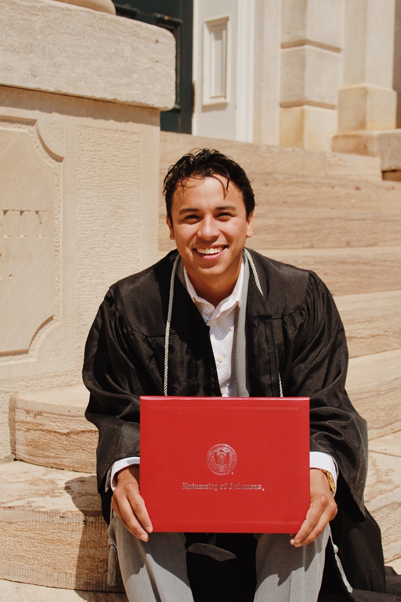 A young man in graduation gown sitting outside on stairs, smiling, holding a red diploma cover with an emblem and the words "Bachelor of Achievements".