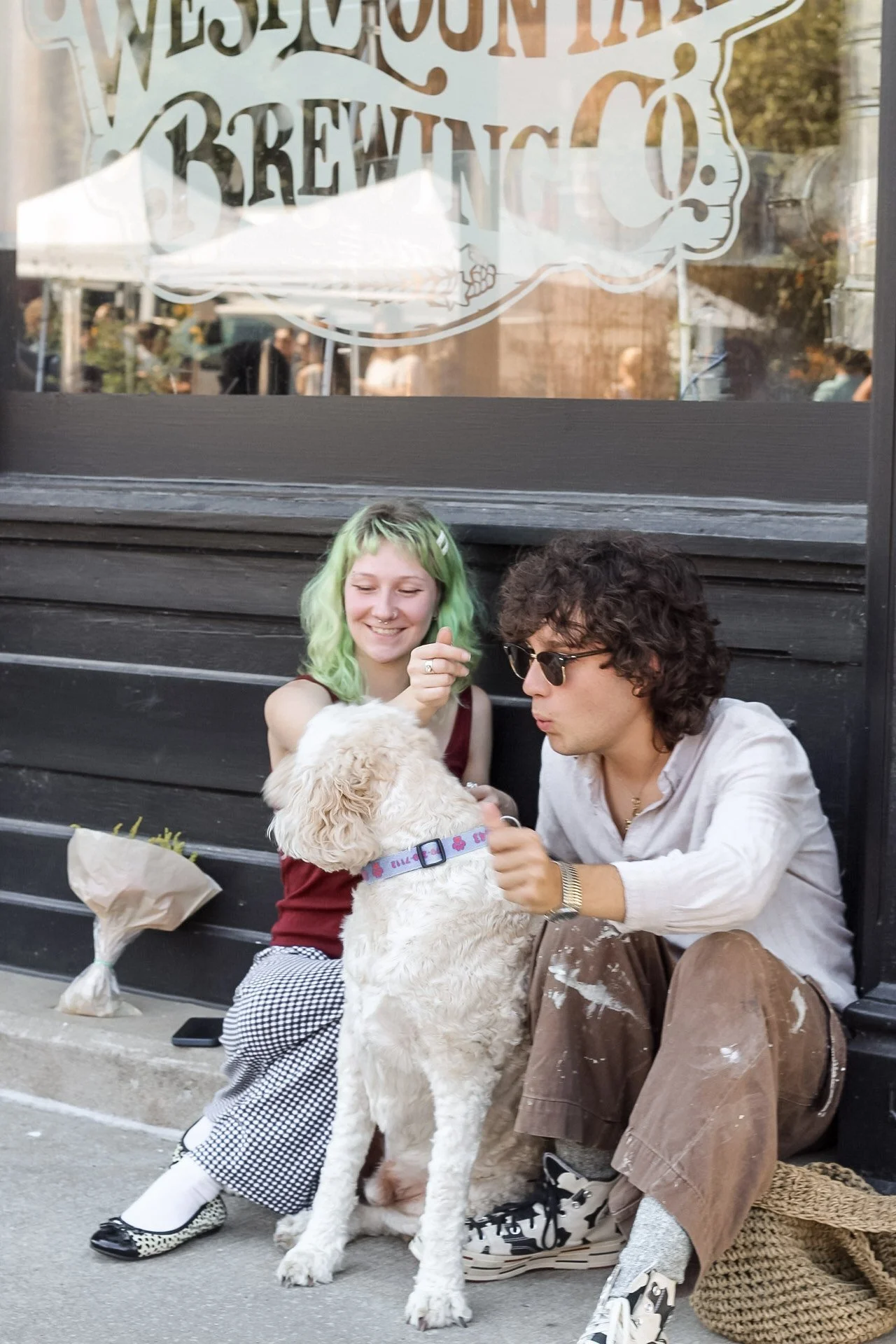 Two young adults, a woman with green hair and a man with curly brown hair wearing sunglasses, sitting on a sidewalk outside a café or restaurant. They are smiling and playing with a large, fluffy, white dog. The woman is wearing a burgundy top and ch