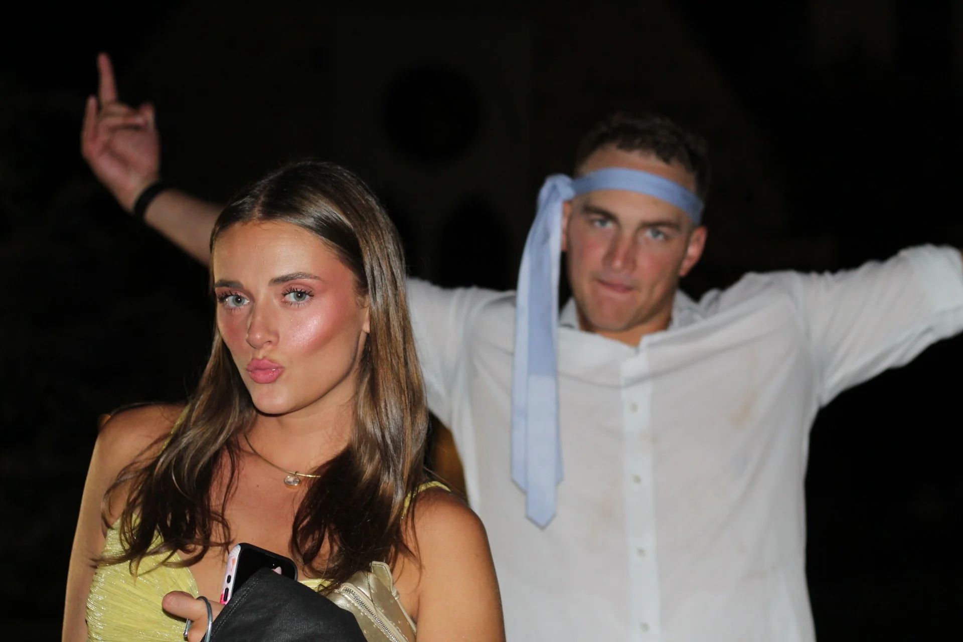 A young woman with long brown hair and a yellow dress making a puckered lips expression, holding a phone and a purse, while a man in a white shirt and blue headband in the background strikes a dance pose with his arms outstretched against a dark back