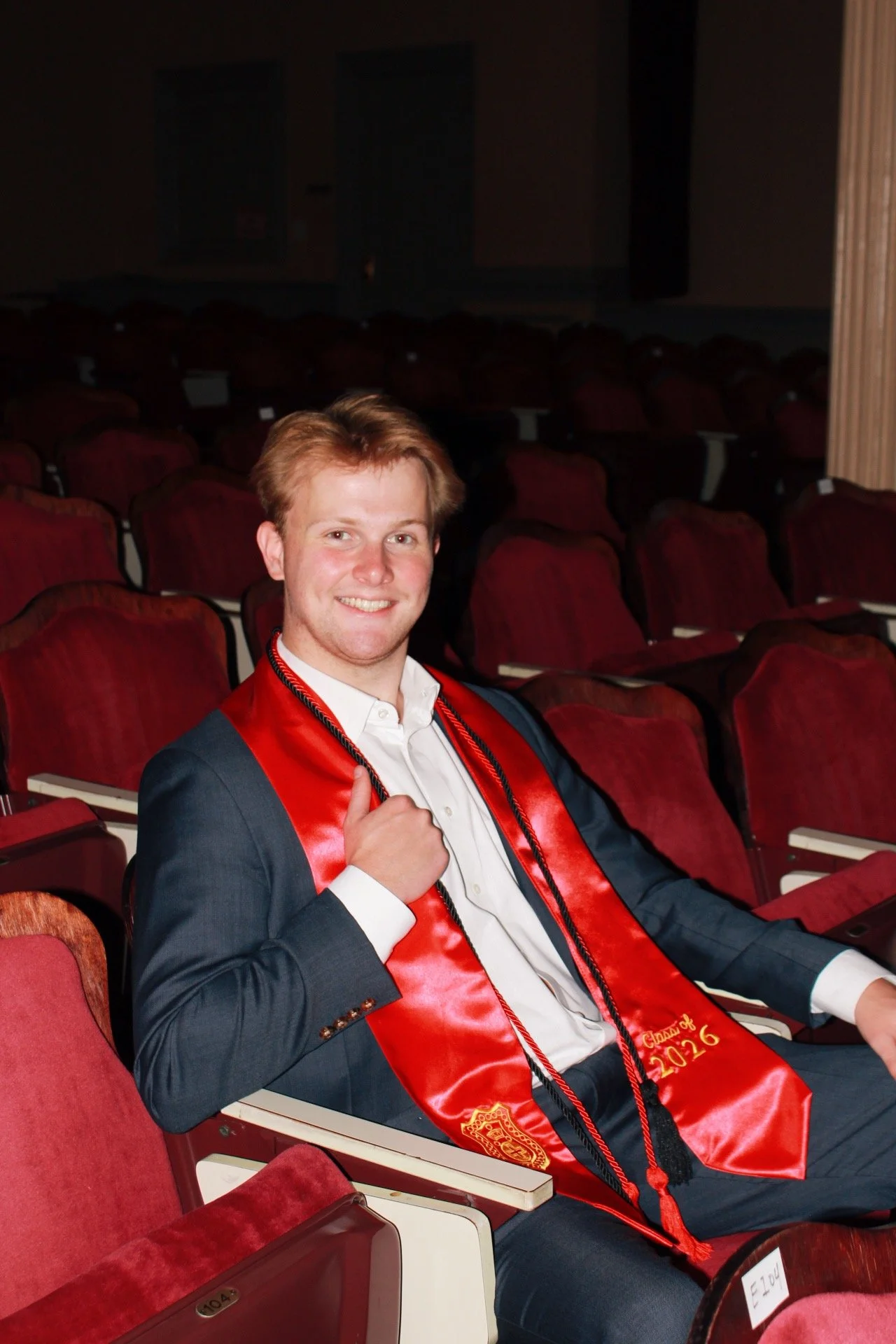 A young man in a graduation cap and gown sitting in an auditorium with red seats, smiling, and wearing a red sash that says 'Class of 2022'.