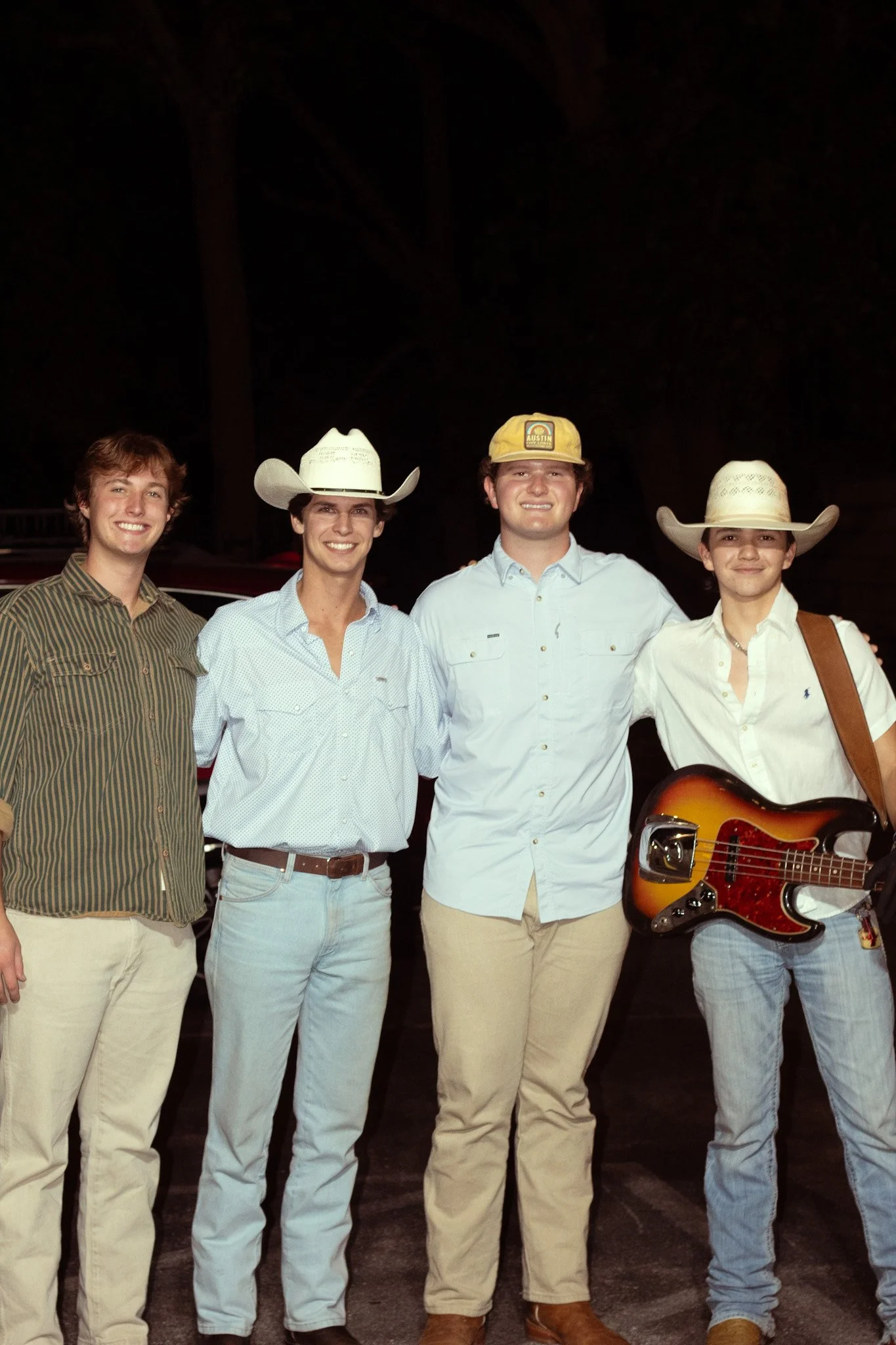 Four young men standing outdoors at night, smiling. The first on the left has curly brown hair and wears a striped brown and green shirt with beige pants. The second wears a white cowboy hat and a light blue shirt. The third wears a yellow cap and a 