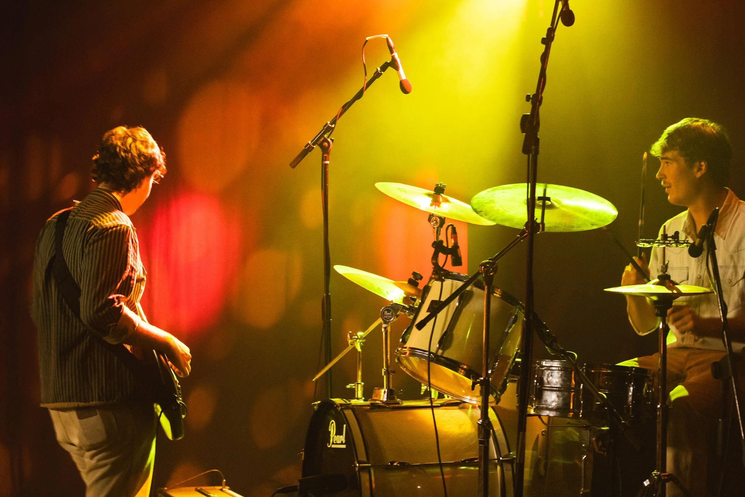 Two musicians perform on stage with drums and microphones under colorful stage lighting.