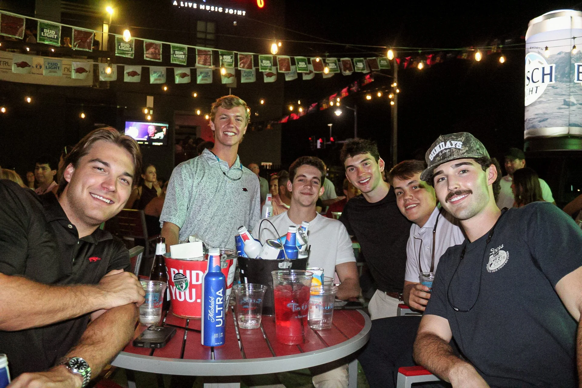 Group of young men smiling and sitting at a table in a lively bar or restaurant with beer bottles, glasses, and drinks on the table. Bright string lights and banners are hanging from the ceiling in the background.