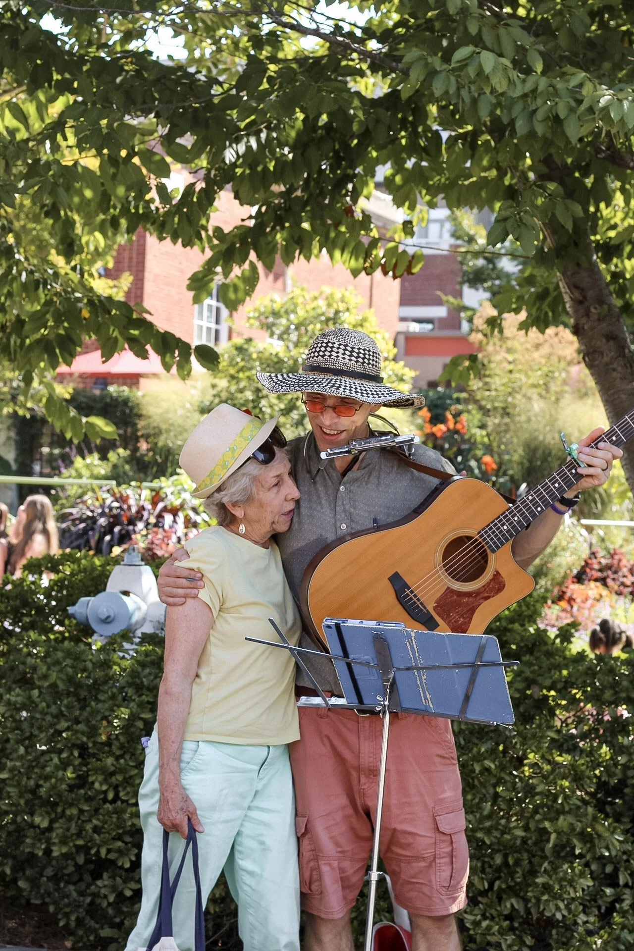 A young man wearing a large sun hat, orange glasses, and a gray shirt is playing guitar and a harmonica; an elderly woman in a yellow shirt and pale blue pants is singing with him under a leafy tree in a park.