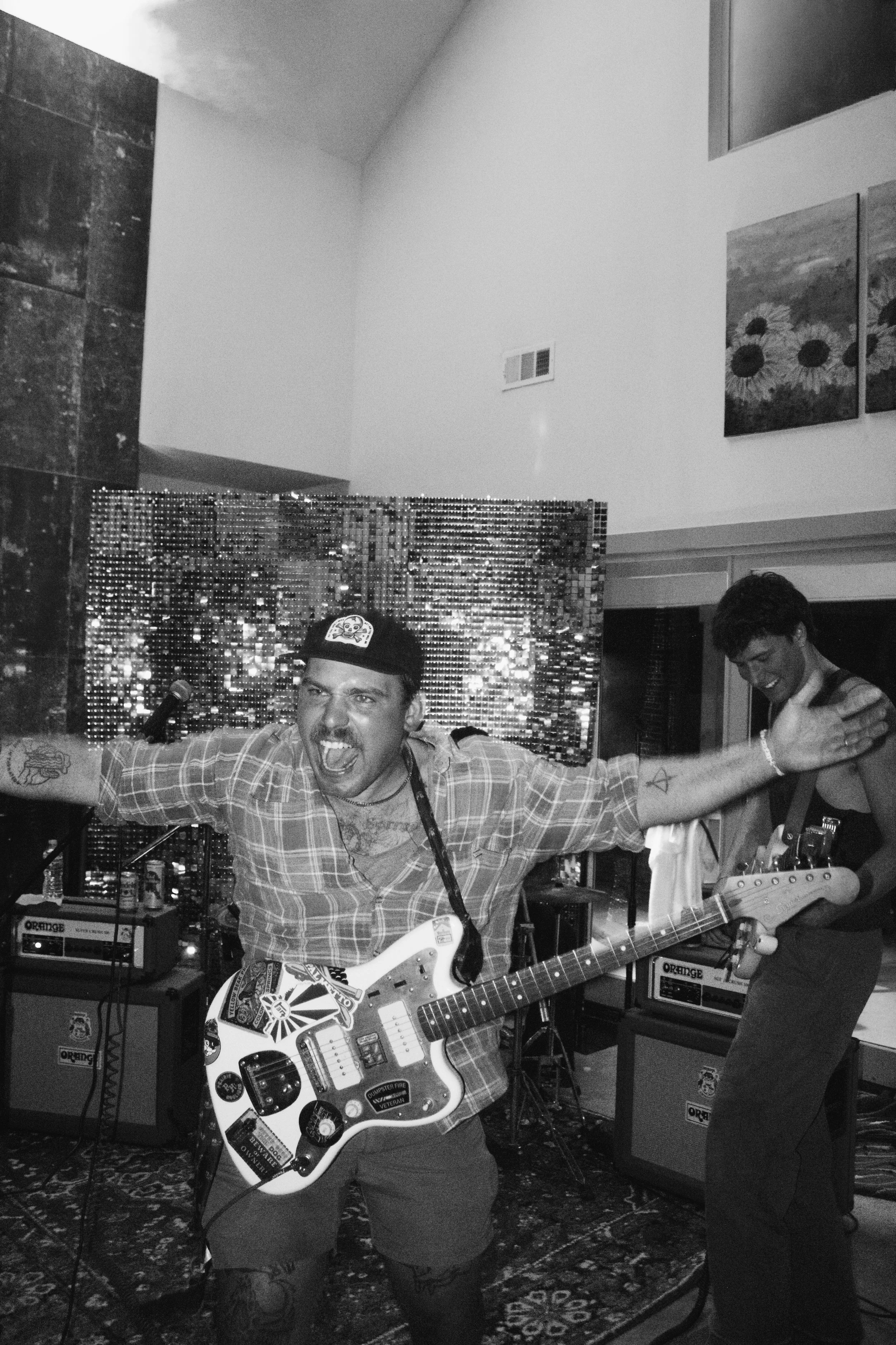 A man playing an electric guitar with a lively expression, wearing a plaid shirt and a baseball cap in a music room. Two other people are visible in the background — one smiling and another partially visible, with music equipment like amplifiers and 