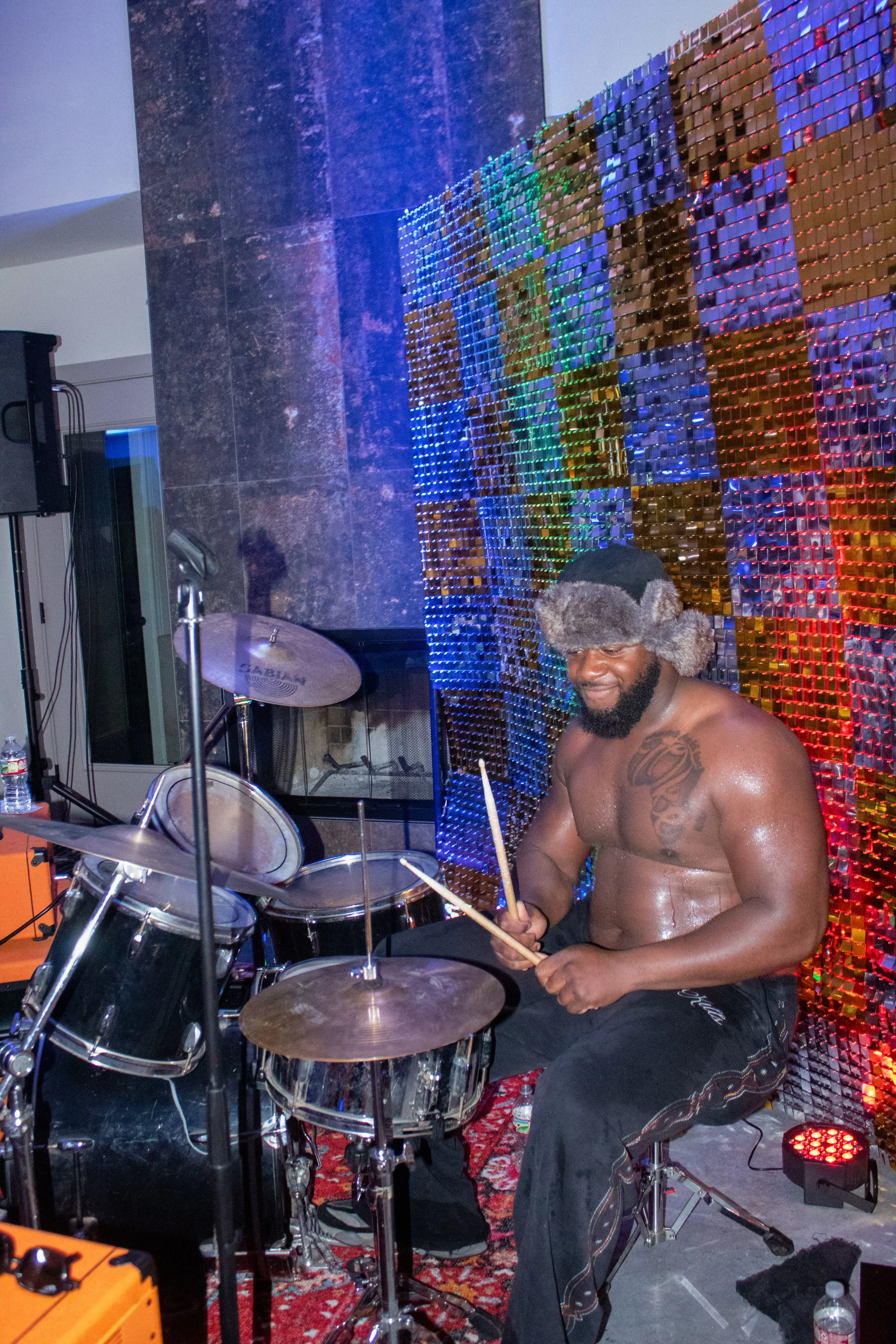 A shirtless man with a beard, wearing a furry winter hat, sitting at a drum set, smiling, with a colorful LED light wall behind him.