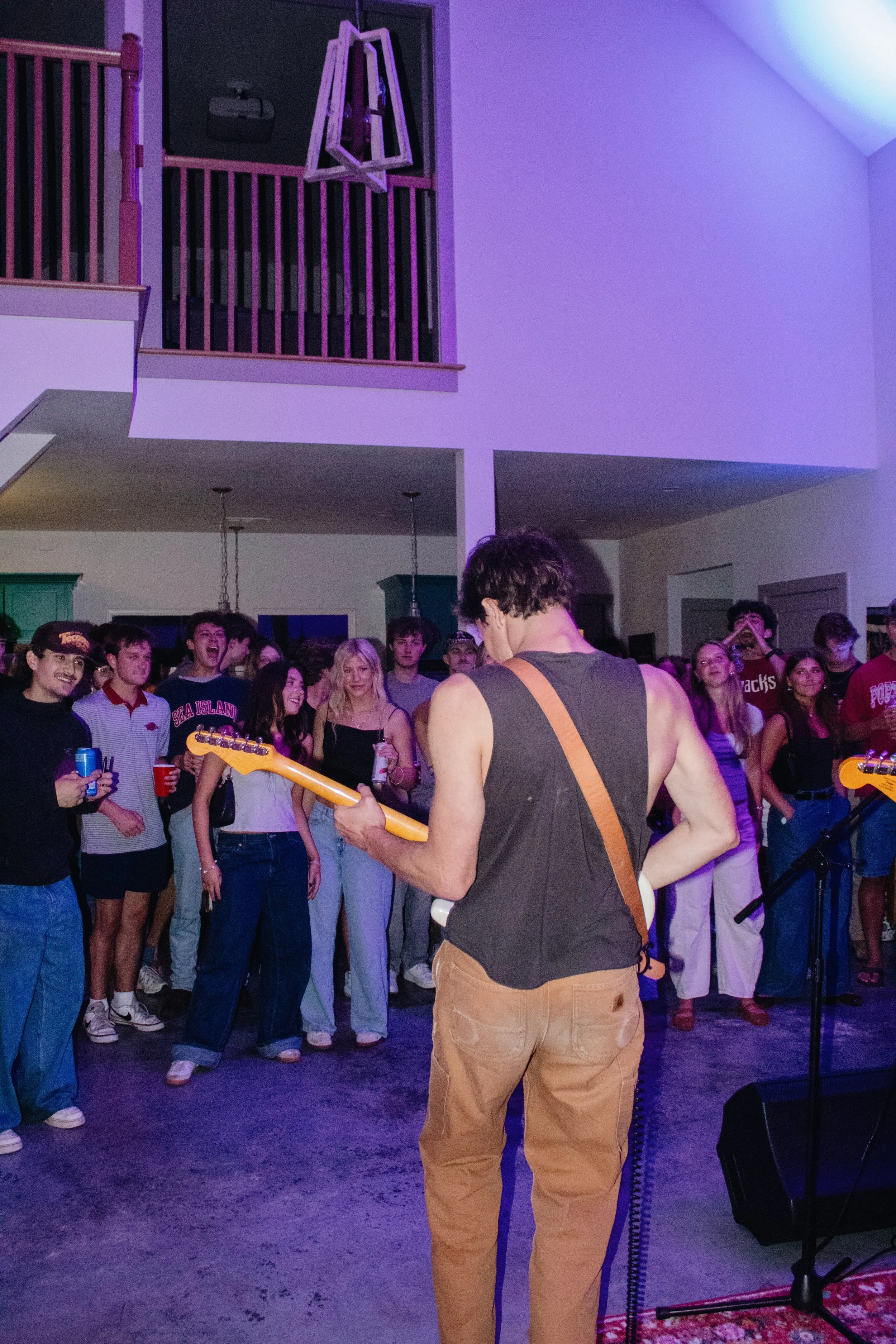 A man in a black tank top and brown pants playing an electric guitar on stage in front of a crowd at a party or concert. The audience consists of young people, some smiling, holding drinks, and watching him perform. The room has a high ceiling and pu