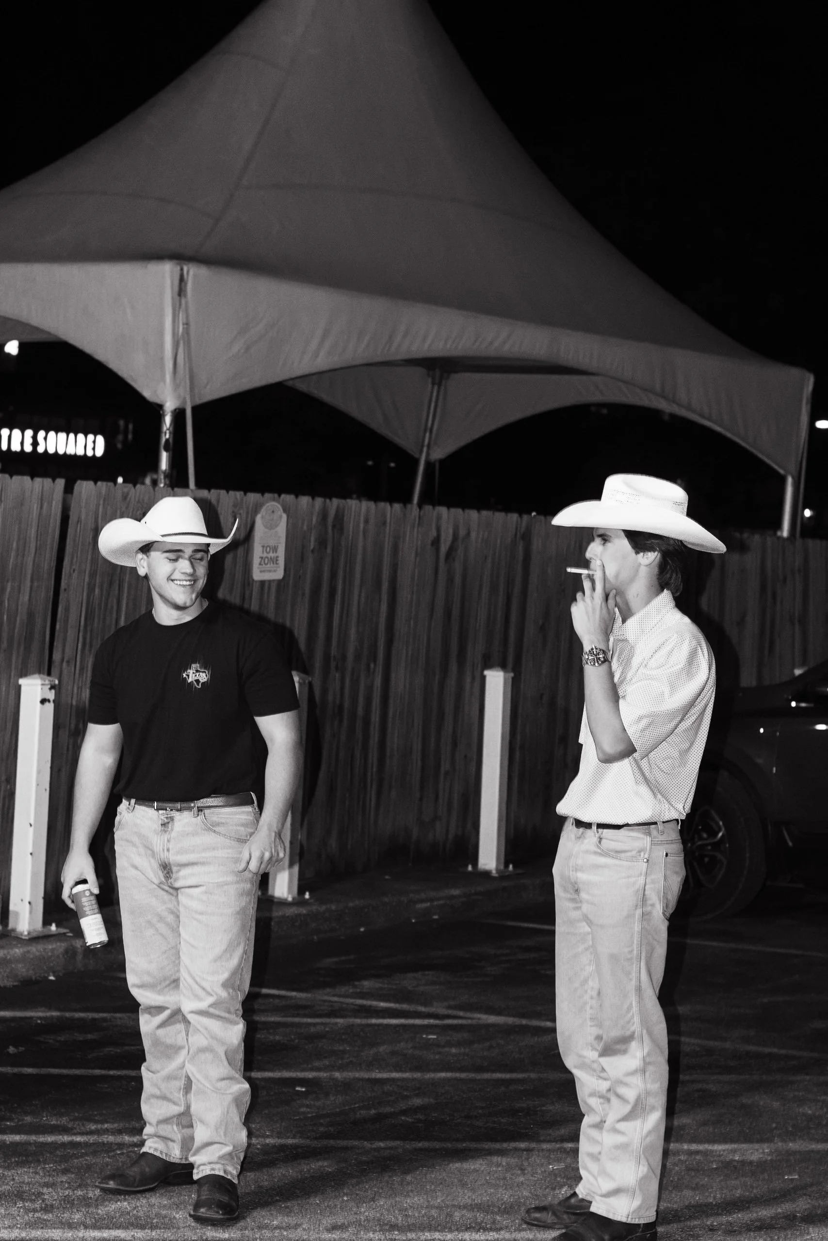 Two young men wearing cowboy hats stand outside at night, one smiling and the other smoking a cigarette. A large tent canopy is overhead, and a wooden fence with a 'Tow Zone' sign is in the background.
