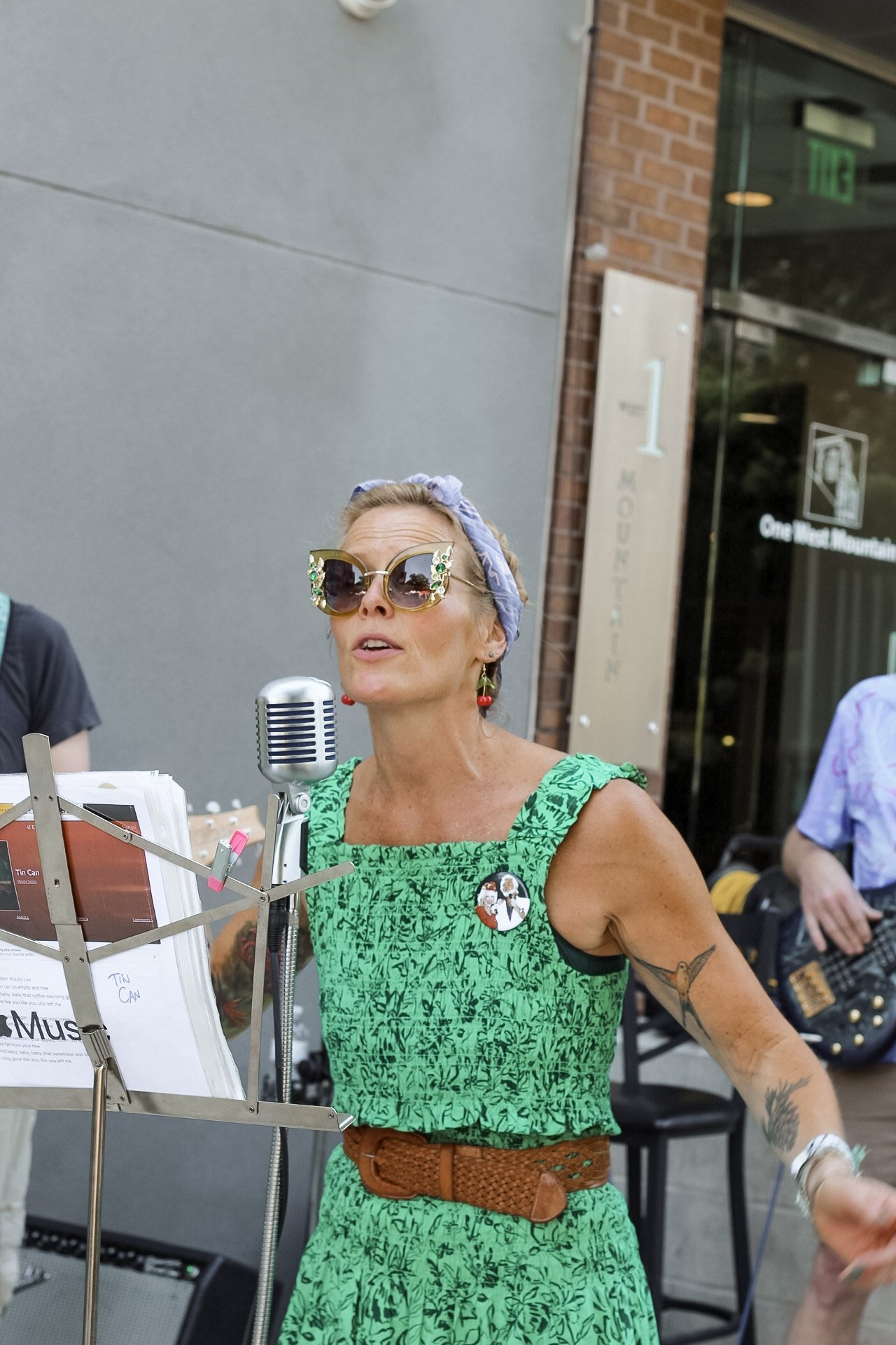 Woman singing into a microphone outdoors, wearing sunglasses, a headscarf, and a green patterned dress with a brown belt, possibly performing at an event.