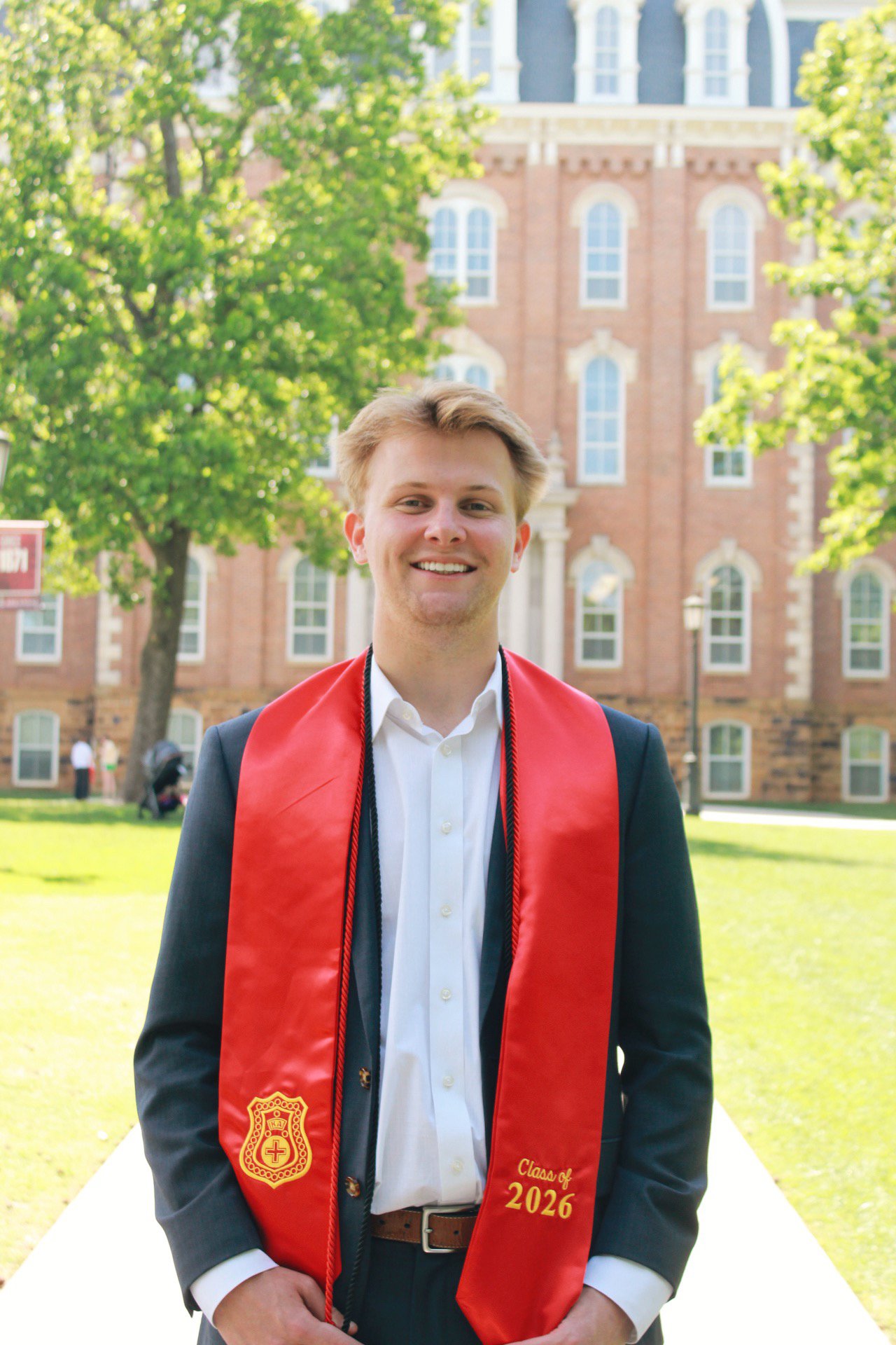 A young man is standing outside on a grassy area, wearing a formal suit, white shirt, and a red graduation stole that says "Class of 2026." He is smiling and has light brown hair. In the background, there are trees, a red brick building with white wi