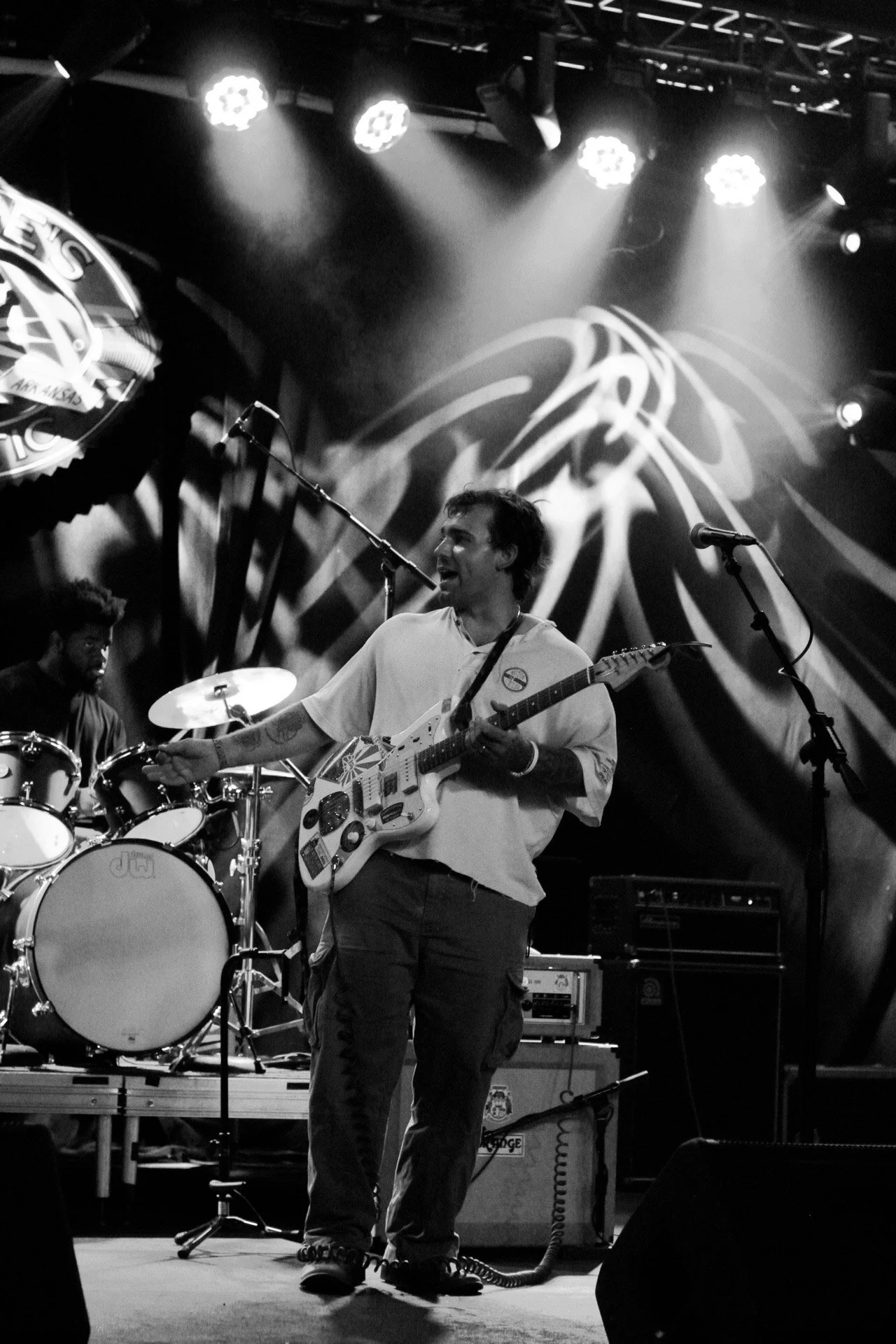 A black and white photo of a band performing on stage. The lead singer is playing an electric guitar, standing in the center, with a drum set and other band members in the background. Stage lights are shining down.