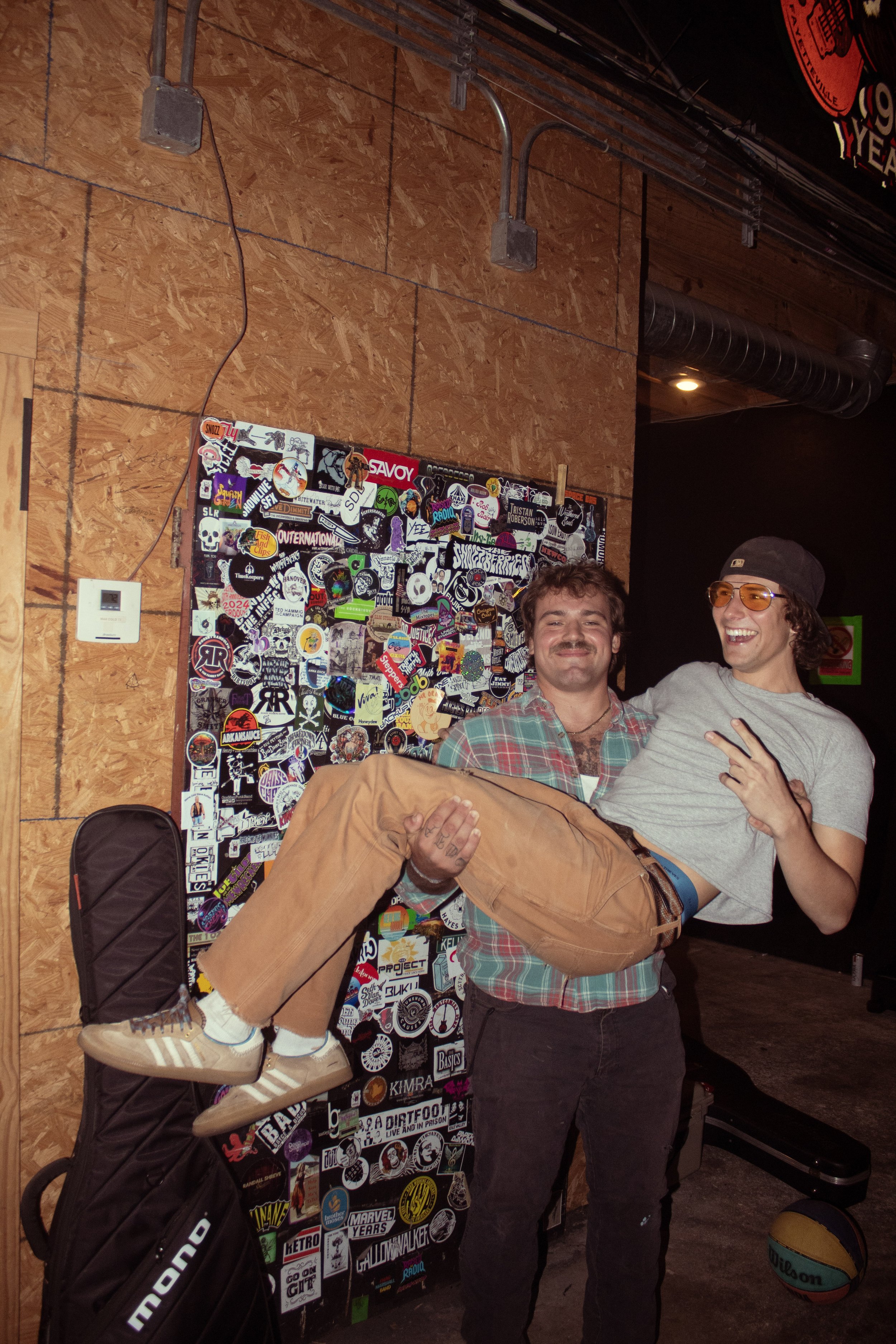 Two men celebrating, one lifting the other, in a room with a wall covered in stickers and a basketball on the floor. The man being lifted is wearing a gray T-shirt and a gray cap, smiling and making a peace sign. The man lifting him is wearing a plai