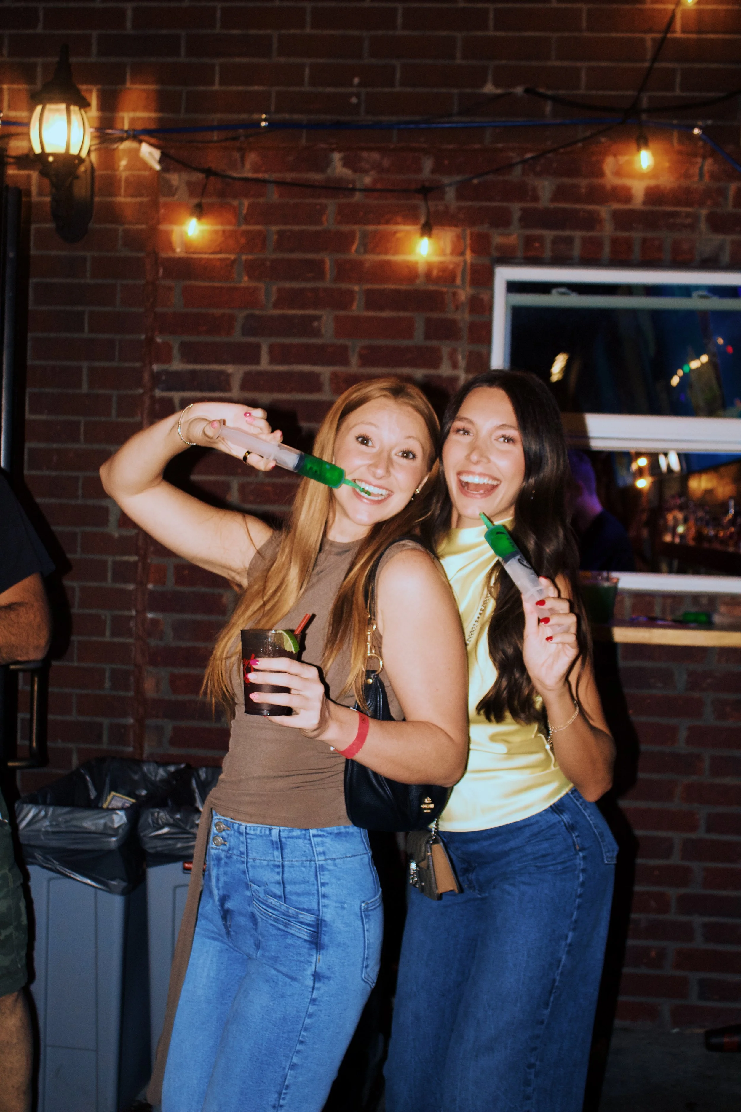 Two women smiling and posing for a photo at a party, holding drinks and shot glasses filled with green liquid, standing in front of a brick wall with string lights.