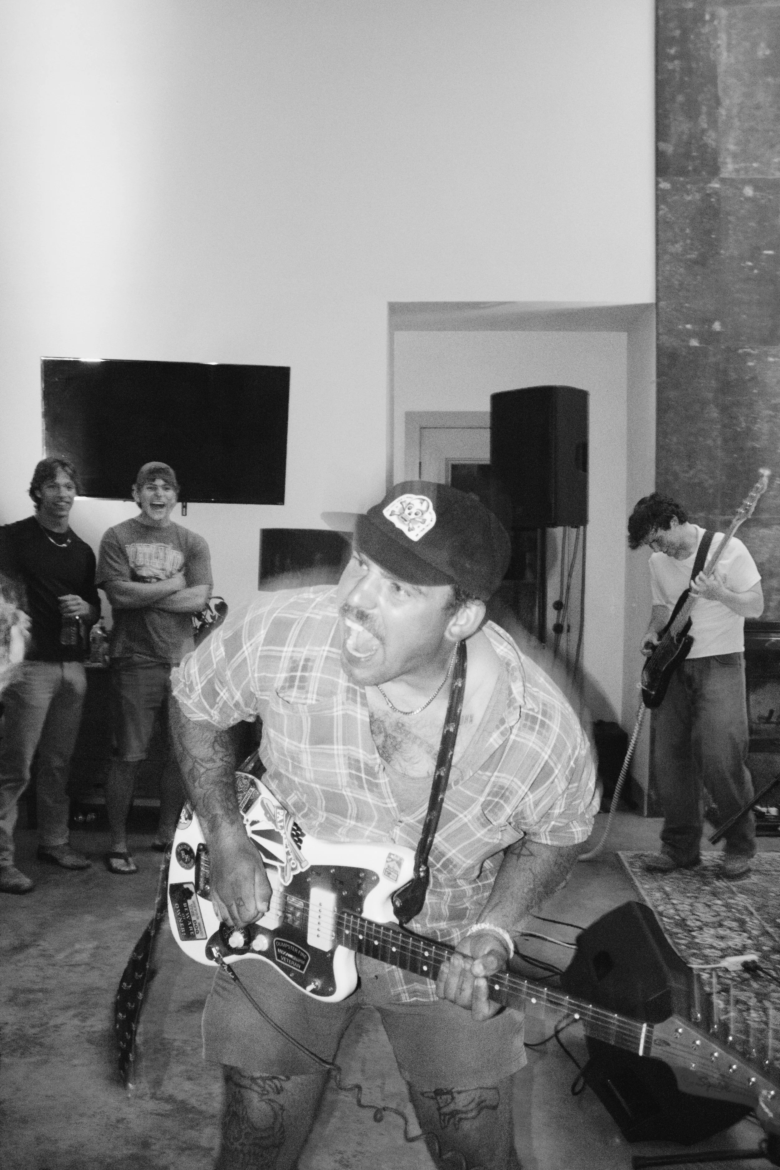 A man with tattoos on his legs playing an electric guitar at an indoor event, with three other men in the background, two of whom are smiling and standing near a TV and speaker.