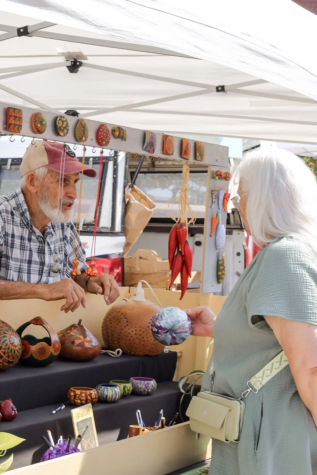 An elderly man and woman at a craft stall, exchanging pottery and decorated gourds, with jewelry and painted objects on display under a white canopy.