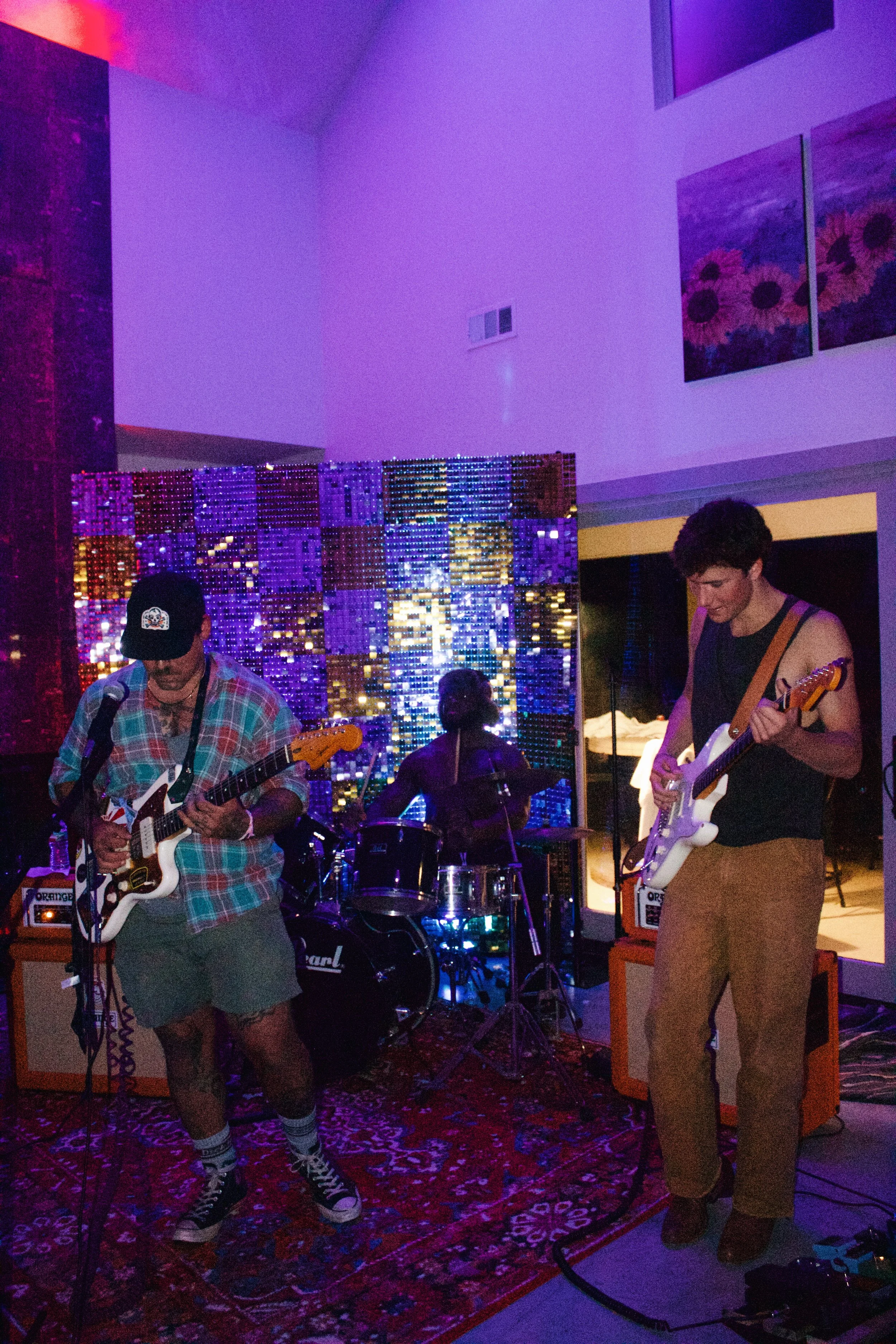 A band of three musicians performing on stage with colorful cityscape lighting in the background, including a guitarist, bassist, and drummer in a dimly lit room.