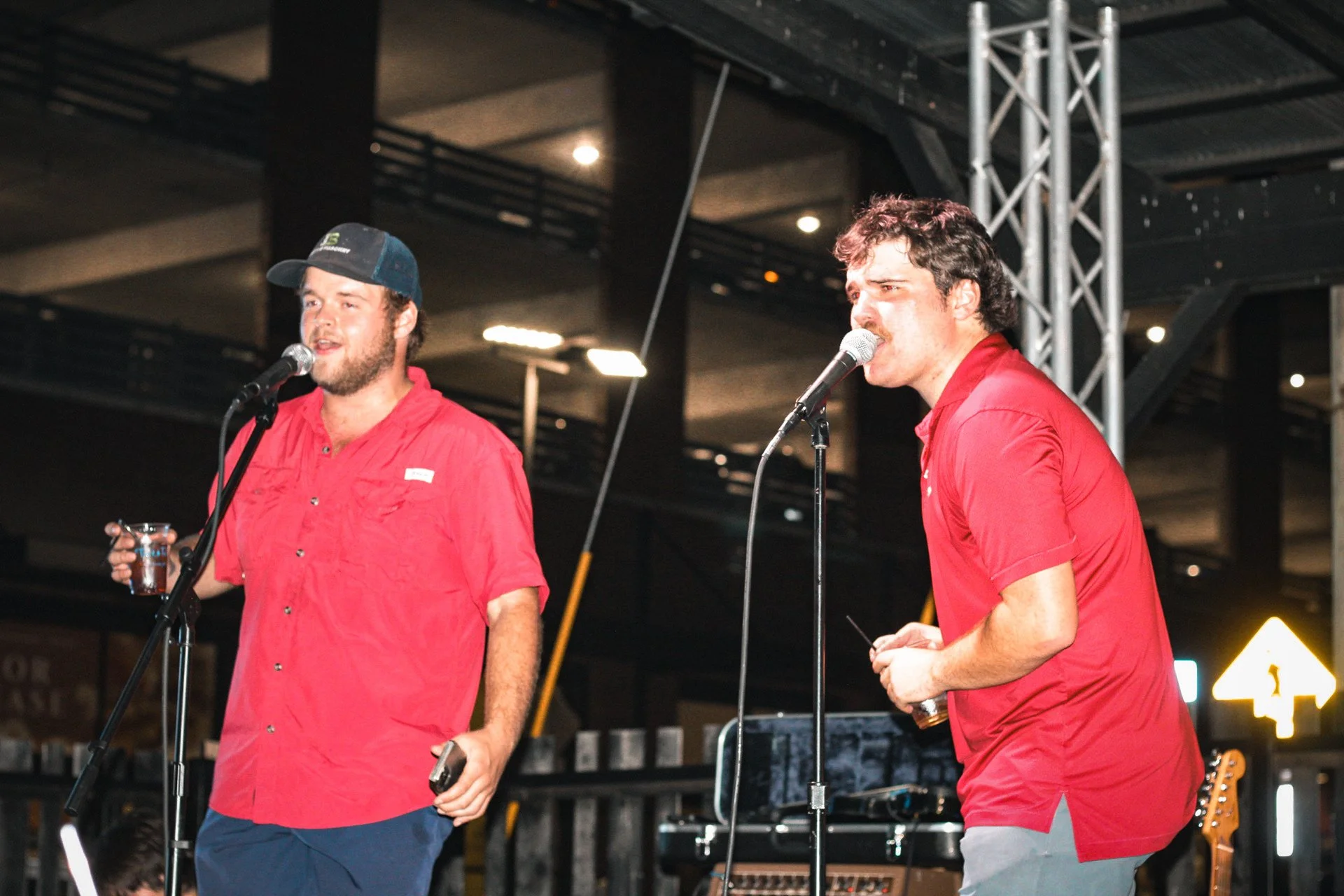 Two men on stage, speaking into microphones. Both wear red shirts. The man on the left has a beard, a cap, and holds a glass of drink. The man on the right has curly hair and a mustache, holding a small object in his hands. Stage equipment and metal 