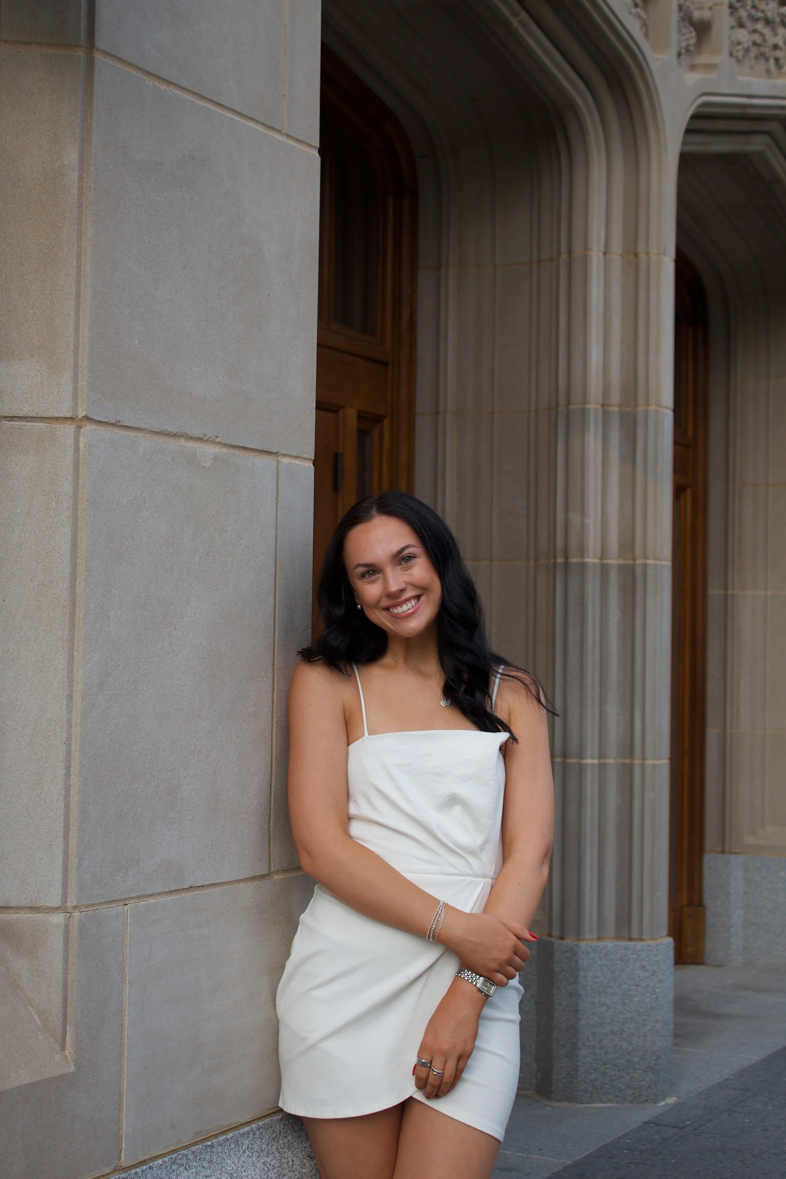 A smiling woman with dark hair wearing a white dress and jewelry leaning against a stone wall outside a building with detailed architectural features.