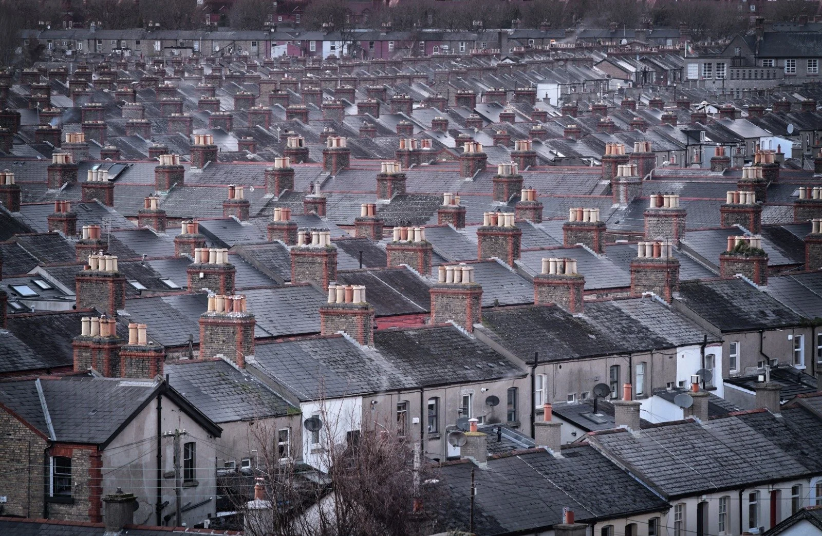 A dense cityscape of rooftops with brick chimneys, some emitting smoke, on cloudy day.