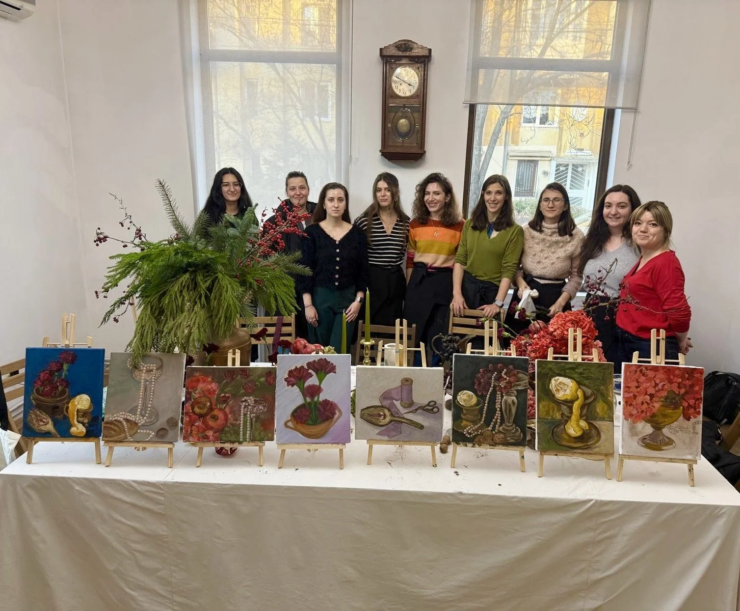 Group of nine women standing behind a table with nine small paintings on easels, floral arrangements, and decorative objects in a room with large windows and a wall clock.