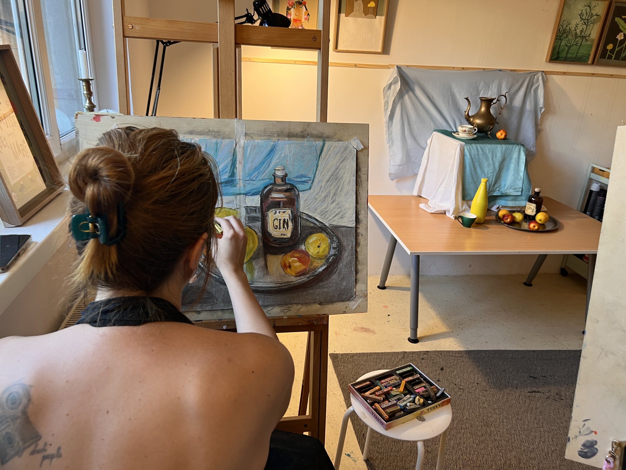 An artist painting a still life of a gin bottle, lemons, and apples on a canvas in a cozy room with a window, various artwork, and a table with more fruits, a yellow bottle, and a teapot.