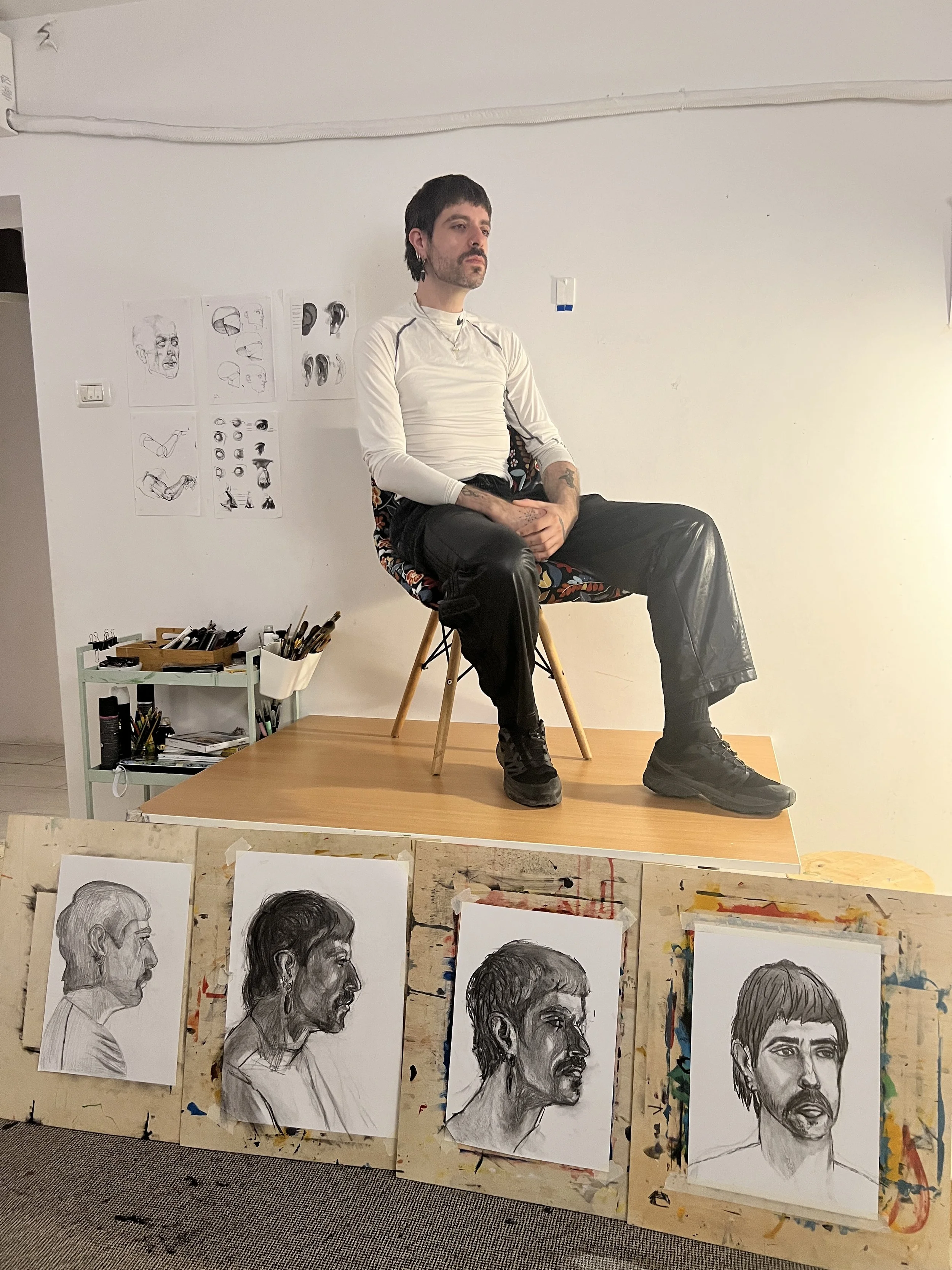 A man sitting on a chair on a table in an art studio, surrounded by drawings and sketches of human profiles and figures.