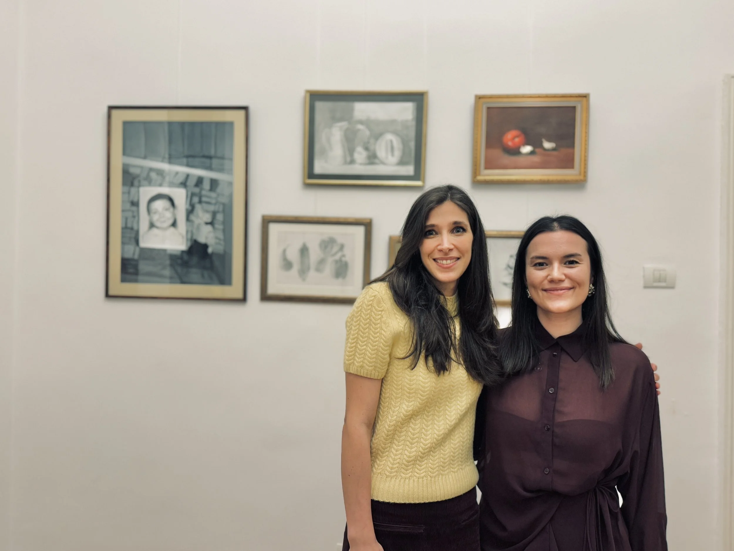 Two women smiling and standing together in front of a white wall with framed artwork, one wearing a yellow sweater and the other a dark purple shirt.