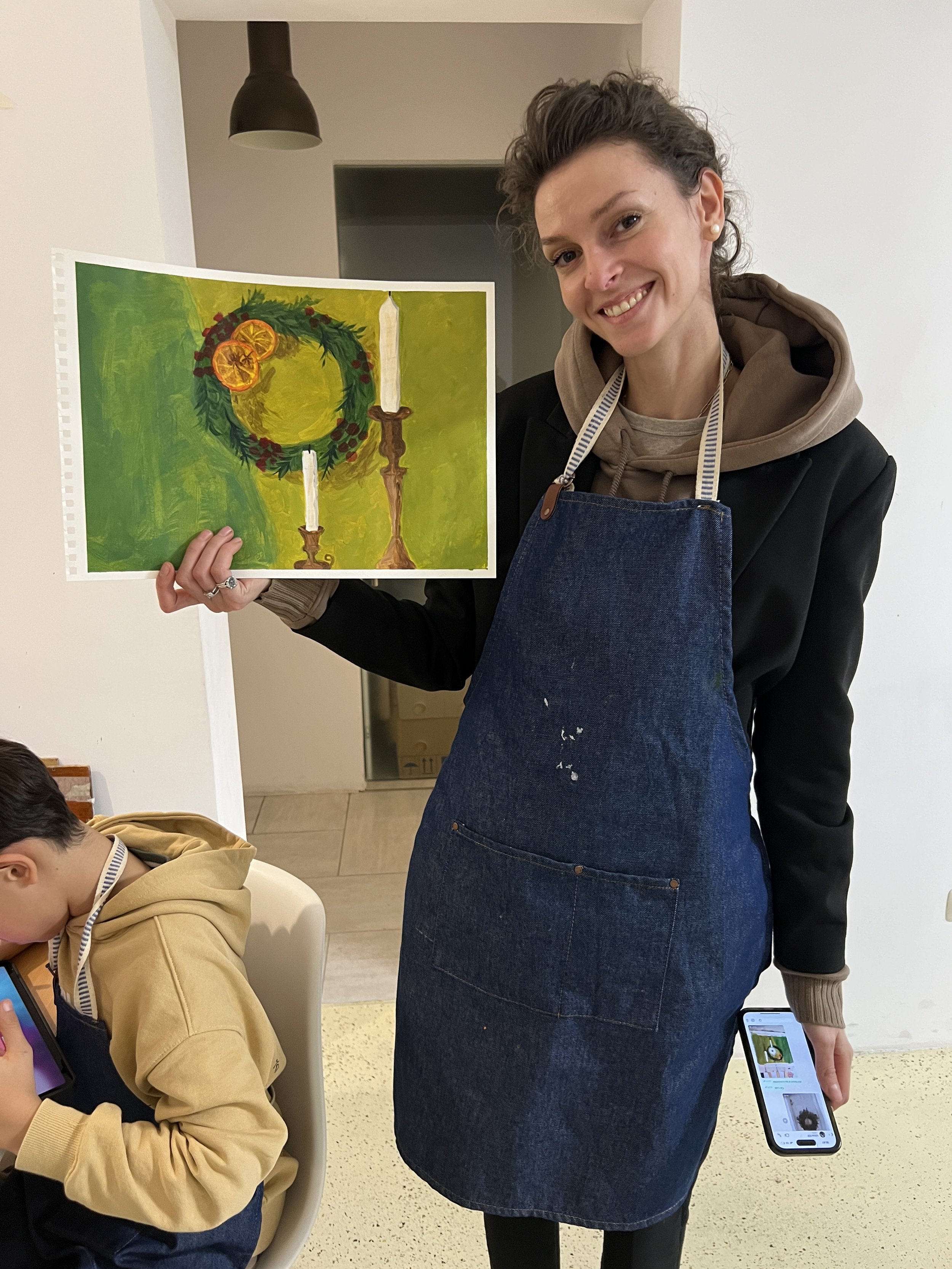 A woman smiling and holding a colorful painting of a holiday scene featuring a wreath with orange slices, a candle, and a red berry garland, with a candle to the right. She is wearing a black jacket, a brown hoodie, and a denim apron. A child in a ye