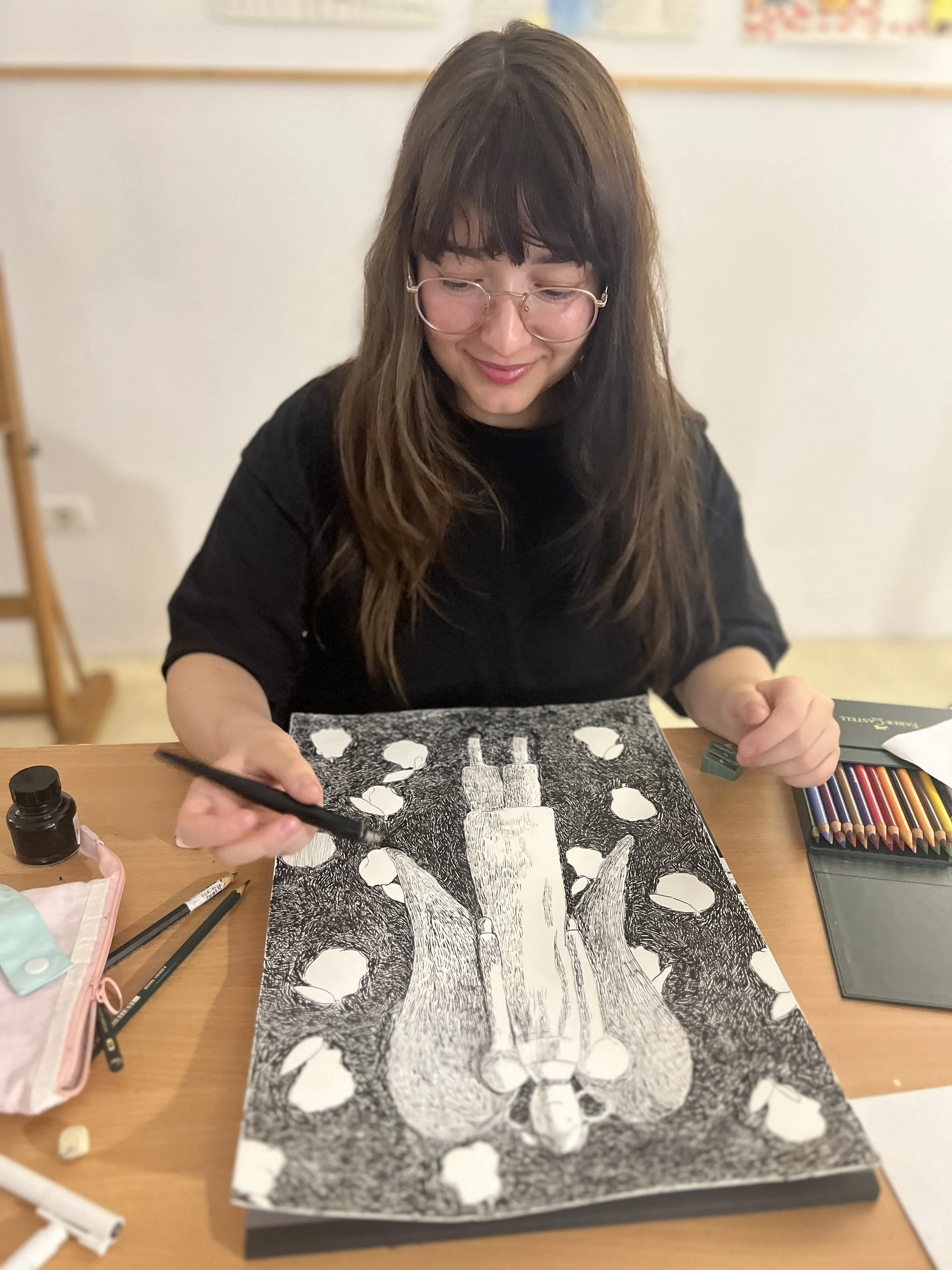 A woman with glasses and long brown hair smiling while drawing a detailed black and white illustration of a dog with her right hand, sitting at a wooden table with art supplies.