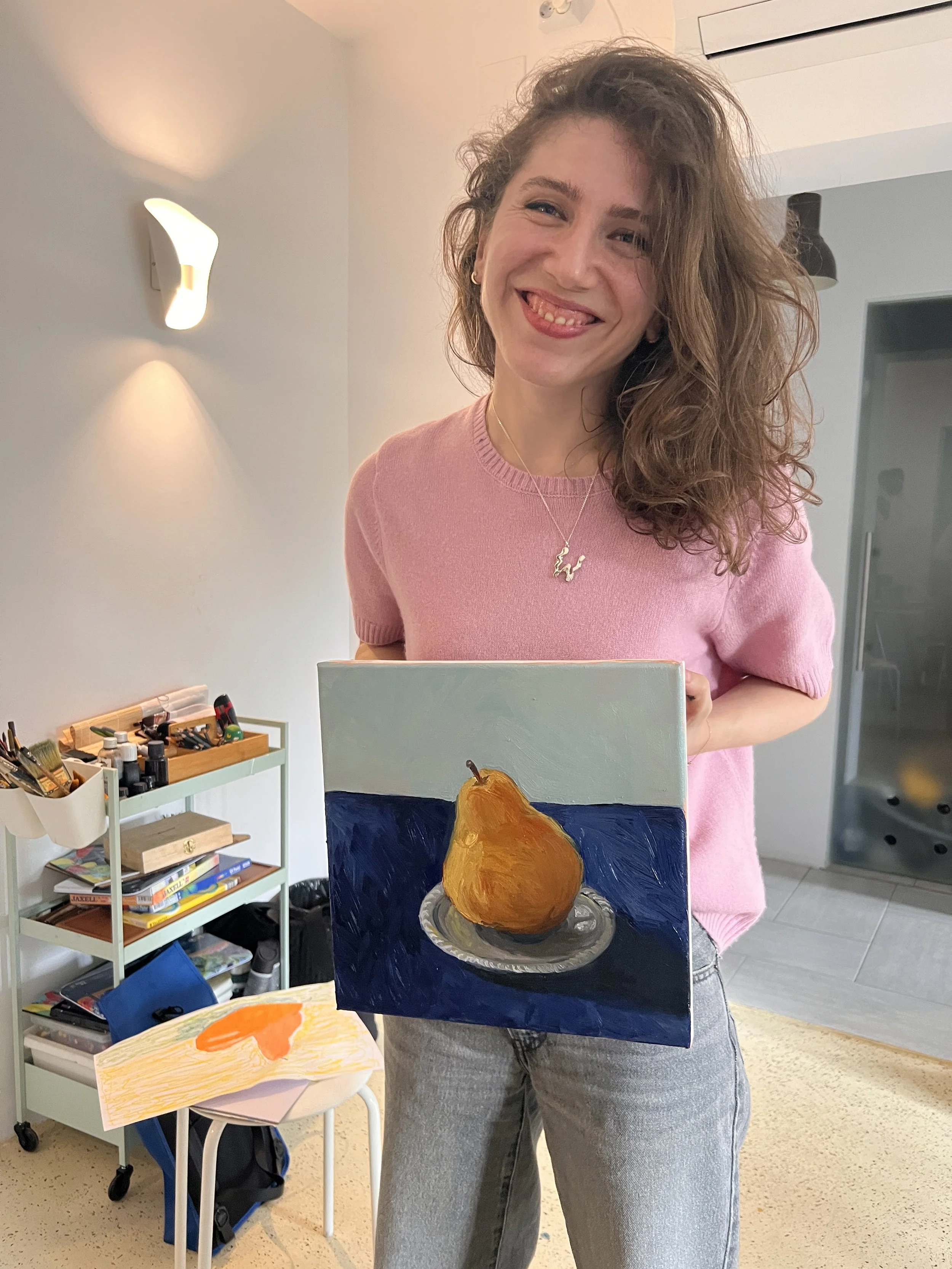 A woman with curly hair in a pink shirt holding a landscape painting of a pear on a plate, smiling in a bright room.