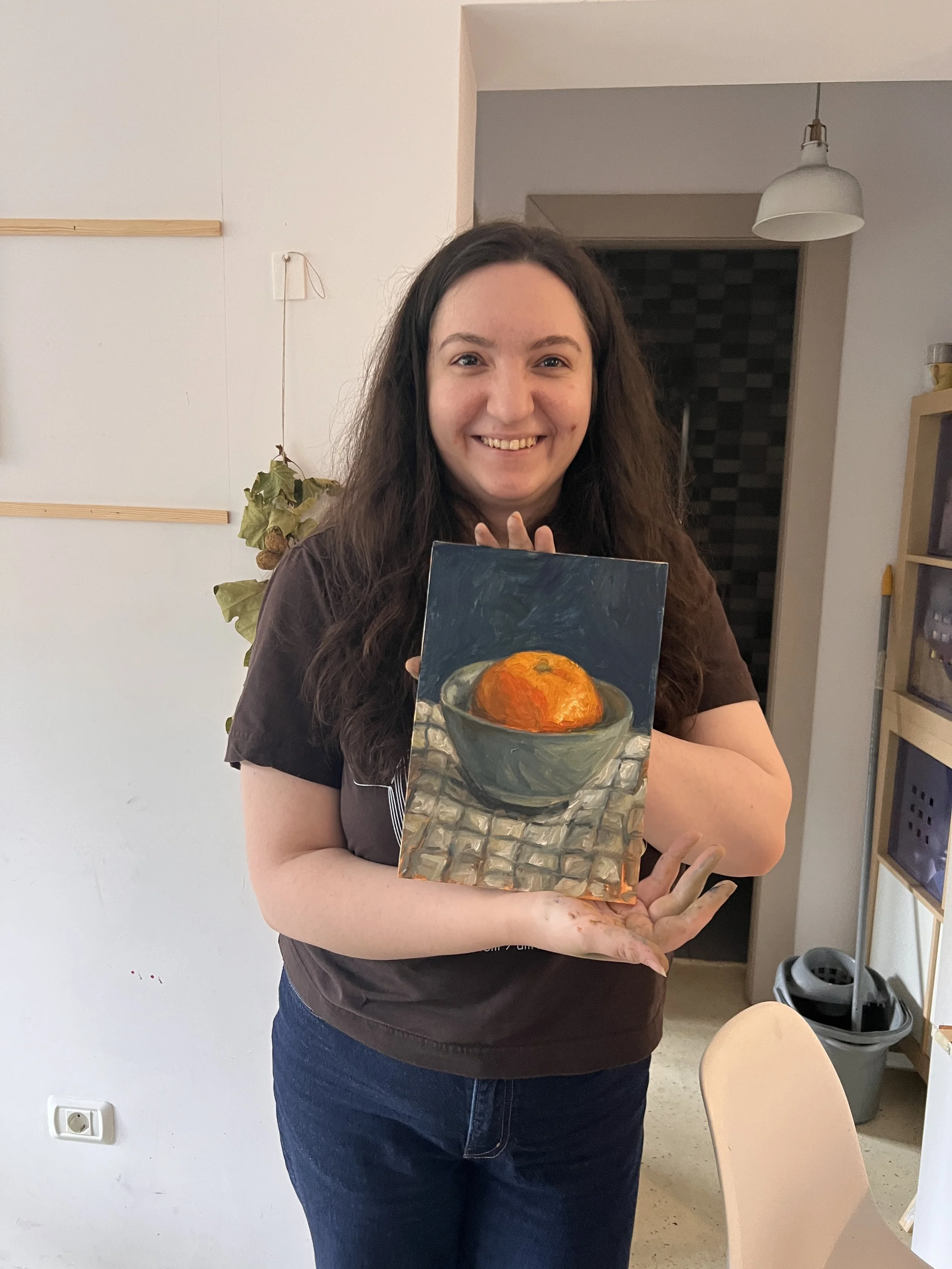 Woman with long dark hair smiling and holding a small painting of a bowl of fruit, possibly an orange.