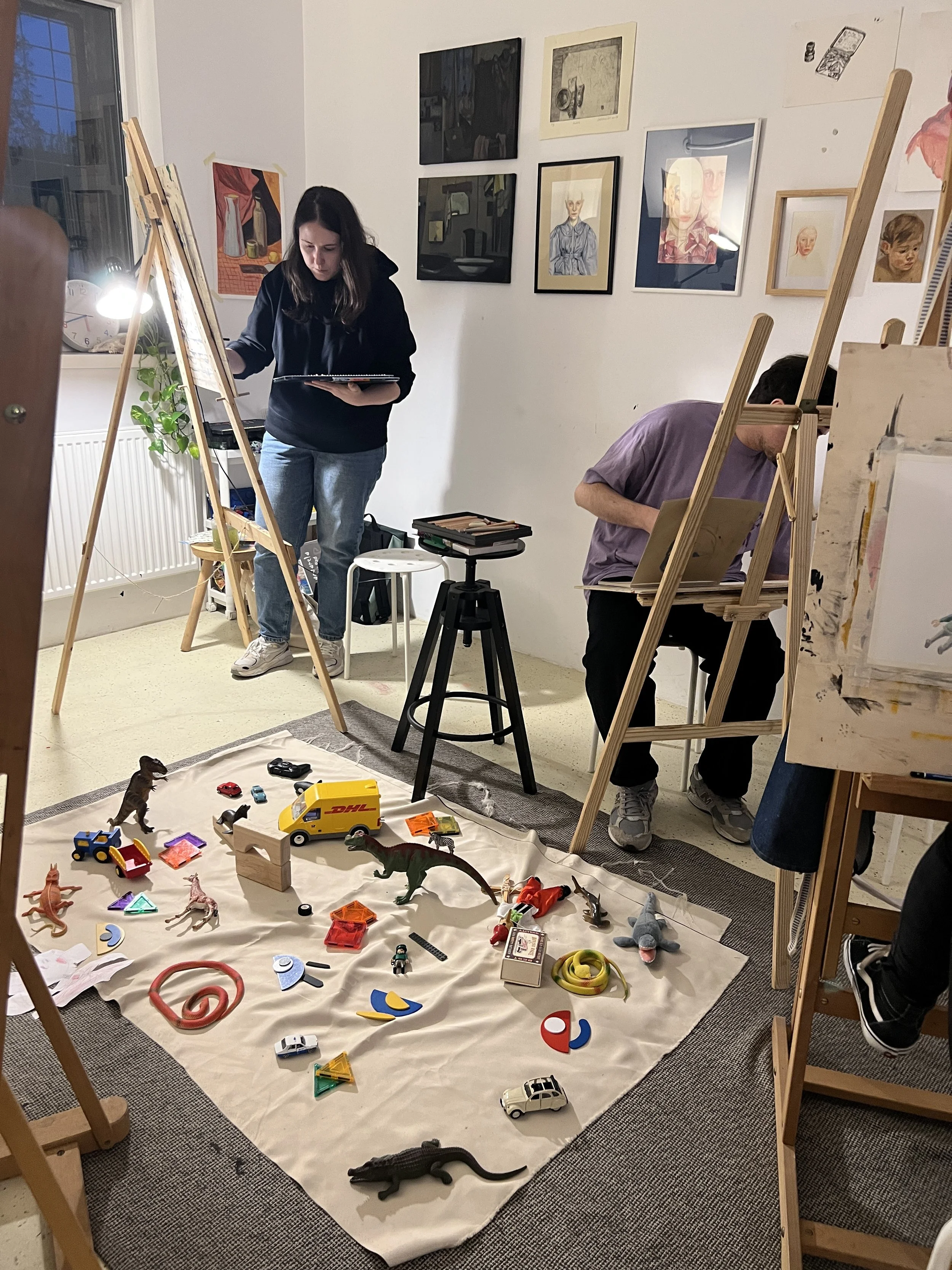 Two artists are working in a studio. A woman stands under a lamp at an easel, while a man sits at his easel. The studio wall is decorated with various framed artworks. The floor is covered with toys, including toy cars, dinosaurs, and geometric puzzl