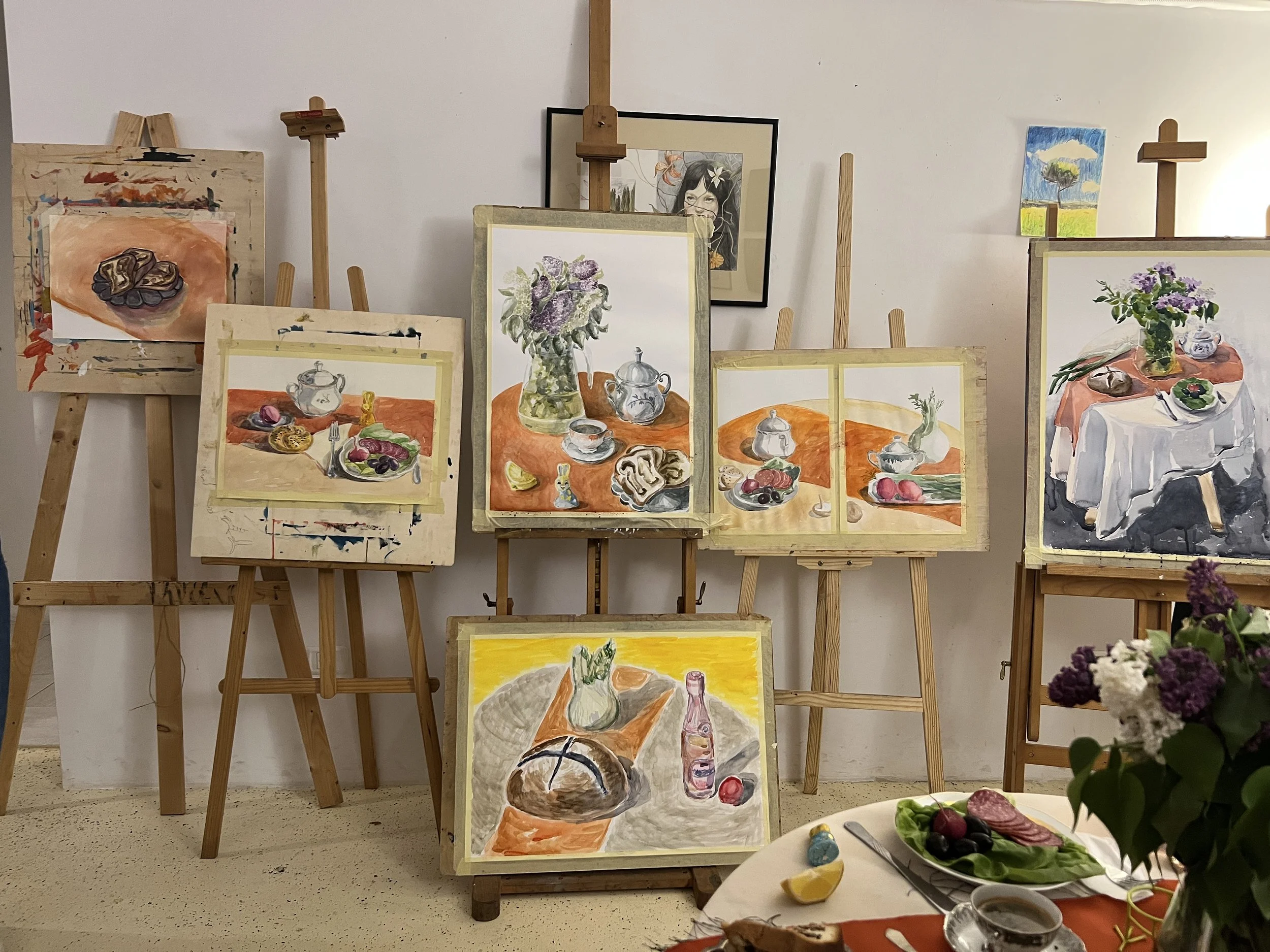 Collection of landscape and still-life paintings on easels and a table in an art studio.