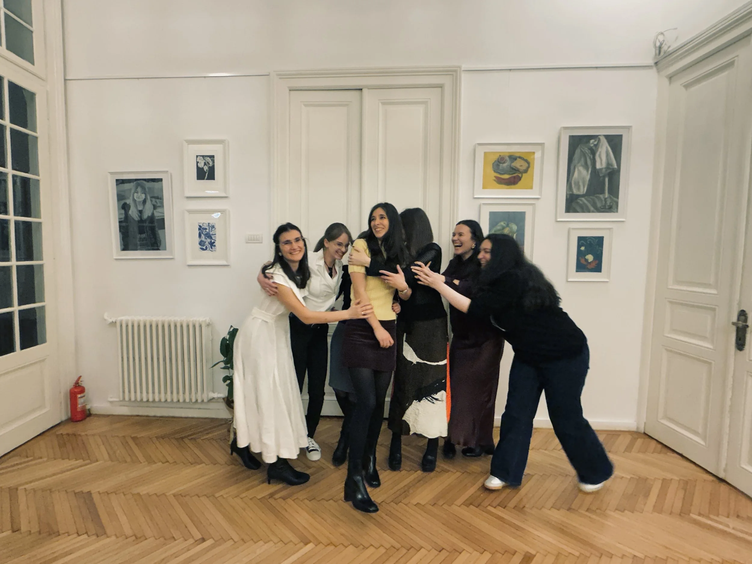 Group of six women hugging and laughing together in an art gallery or living room with framed pictures on the wall.