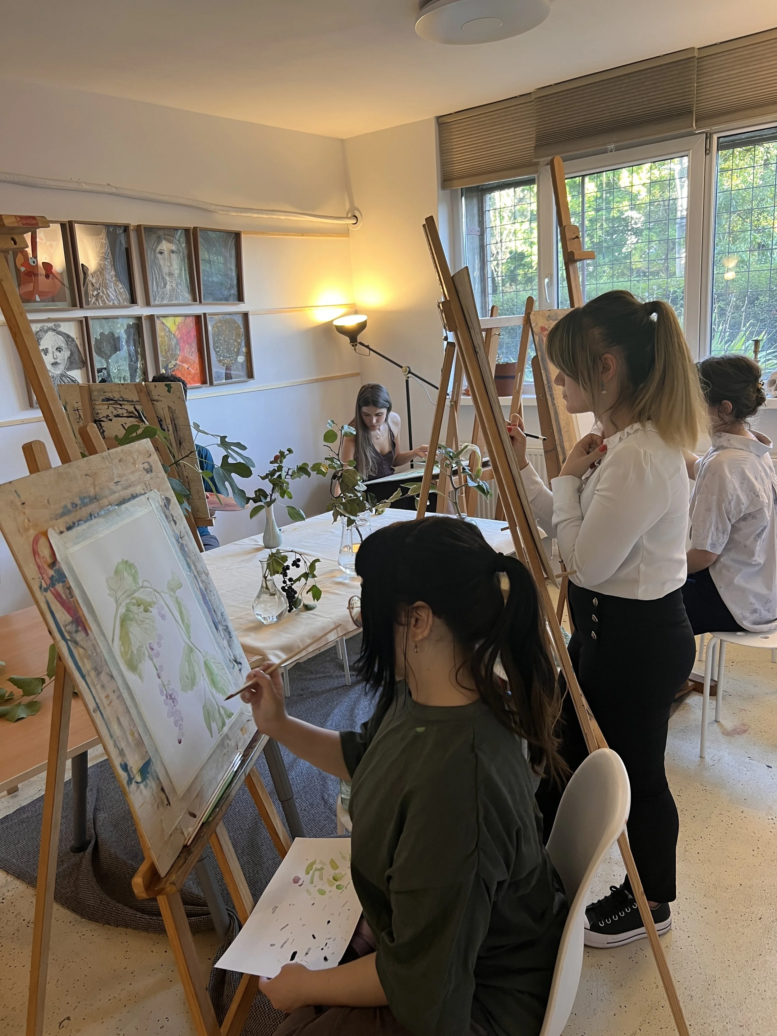 Young women painting on canvases in an art class. One is standing and observing, while another is seated and working on a watercolor. The room has paintings on the wall, plants, and natural light coming from the window.
