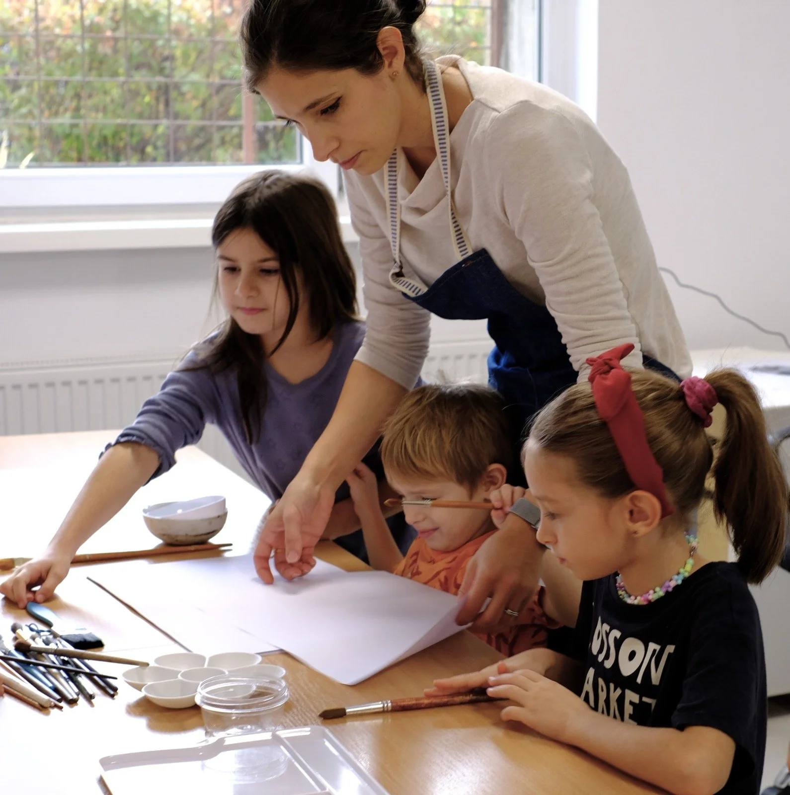 A woman and three children gathered around a table engaged in an art activity, with brushes and watercolor supplies on the table.