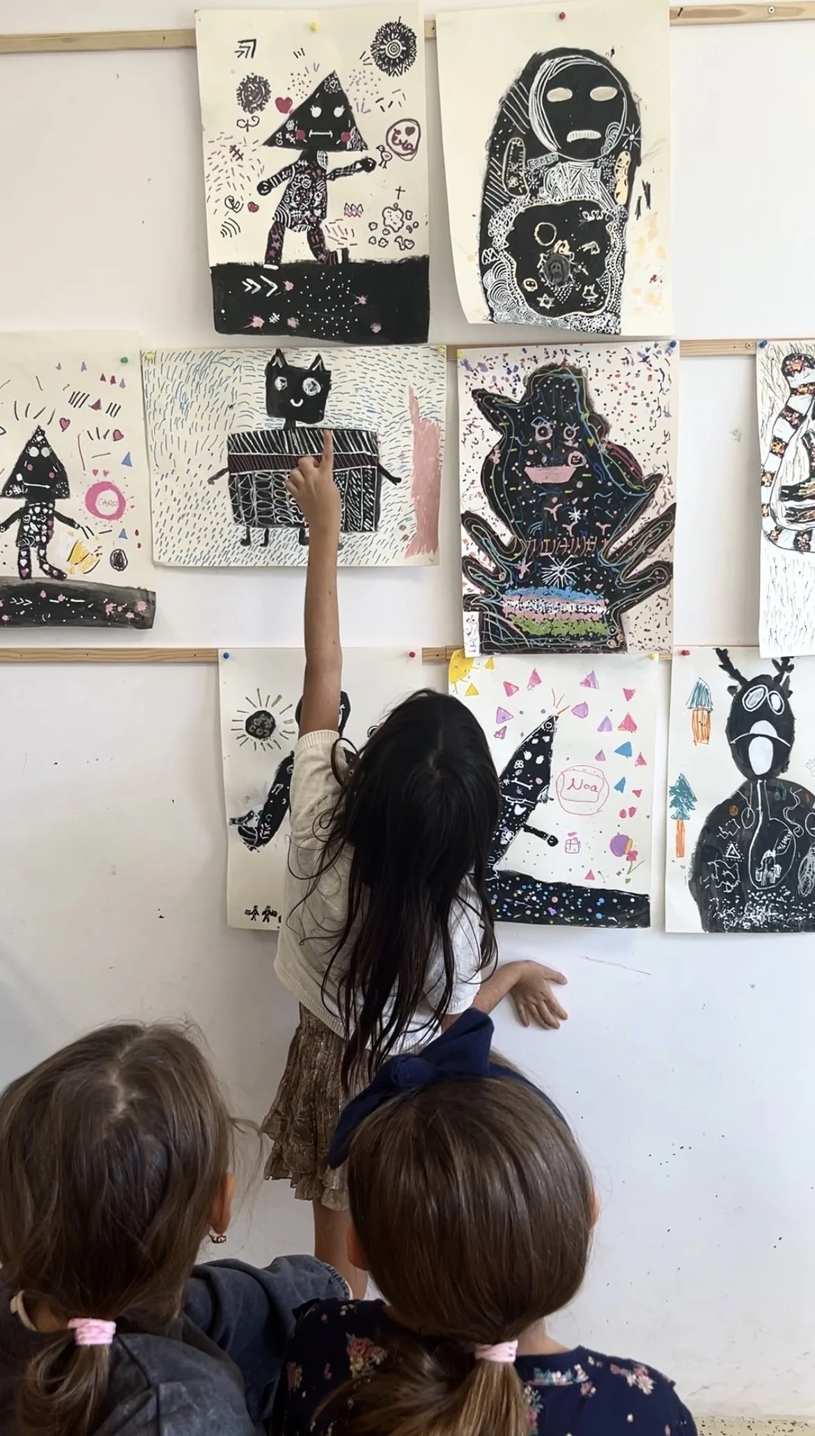 Children viewing colorful abstract artwork and drawings of animals and characters displayed on a wall in a classroom or art gallery.