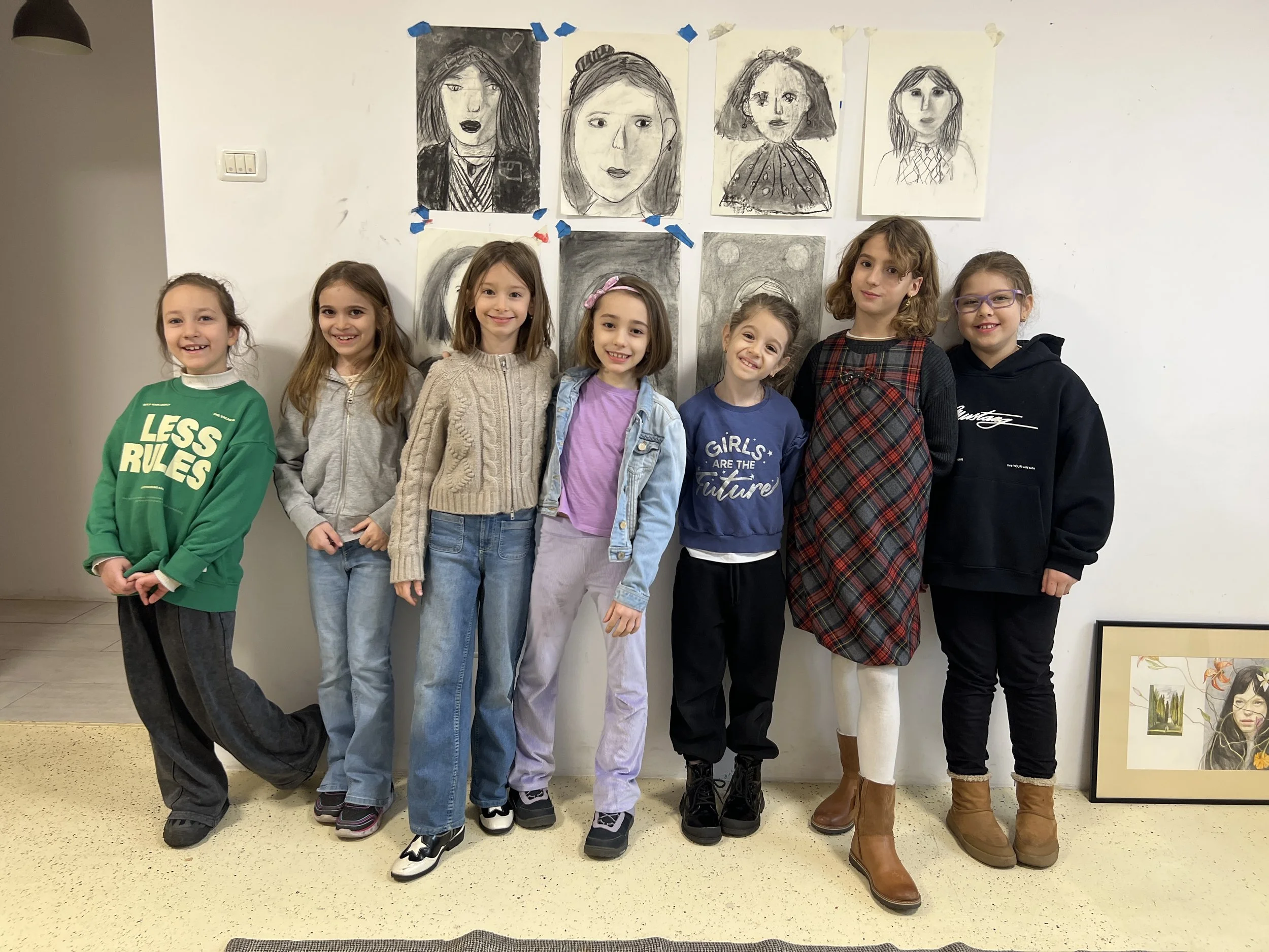 Group of eight young girls standing in front of a white wall with drawing and paintings of faces displayed above them. They are smiling and posing for the photo.