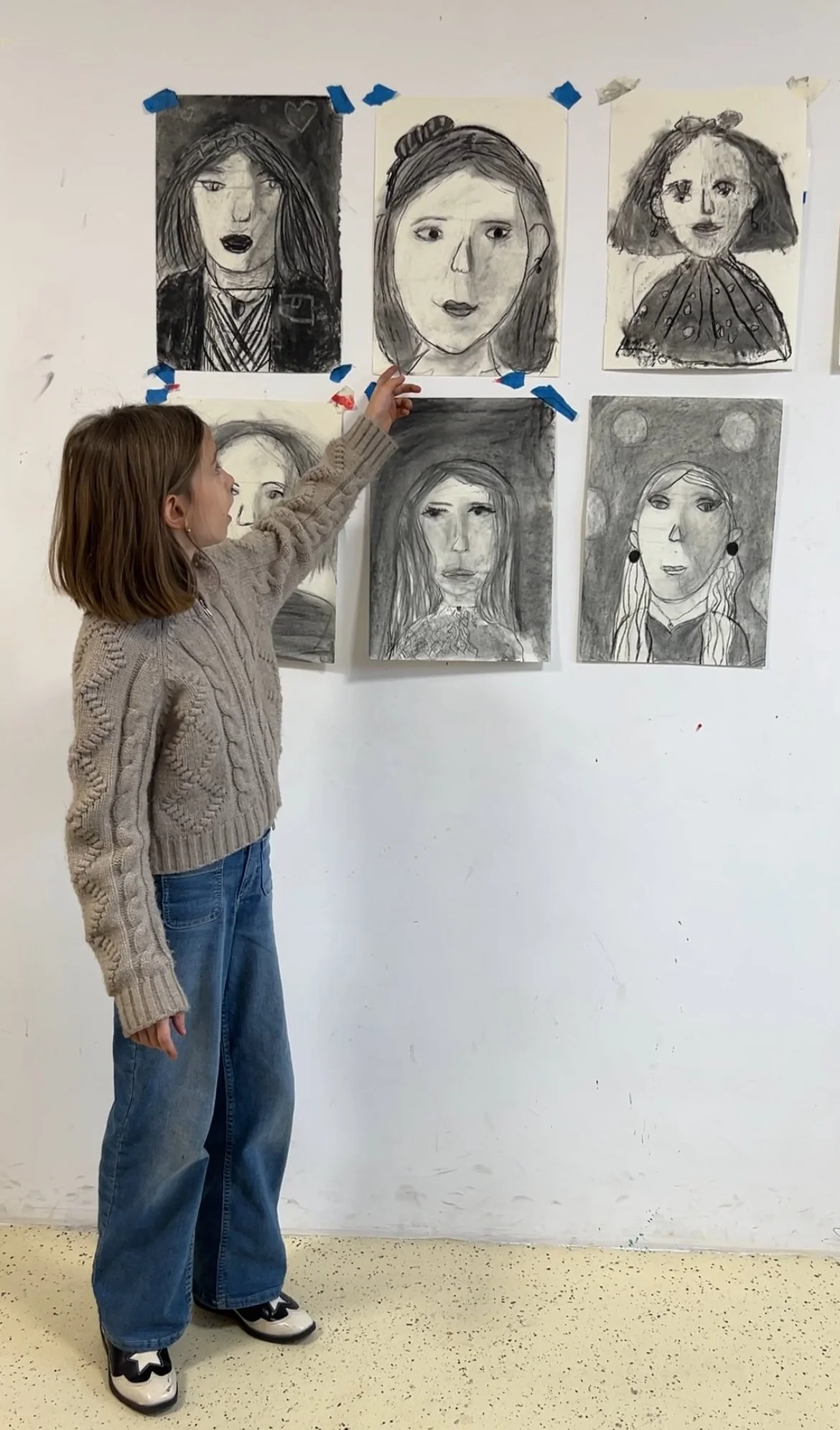 A young girl in a beige sweater and blue jeans is pointing at a portrait on a wall. The wall displays seven black and white portrait drawings of women, taped with blue and red tape.