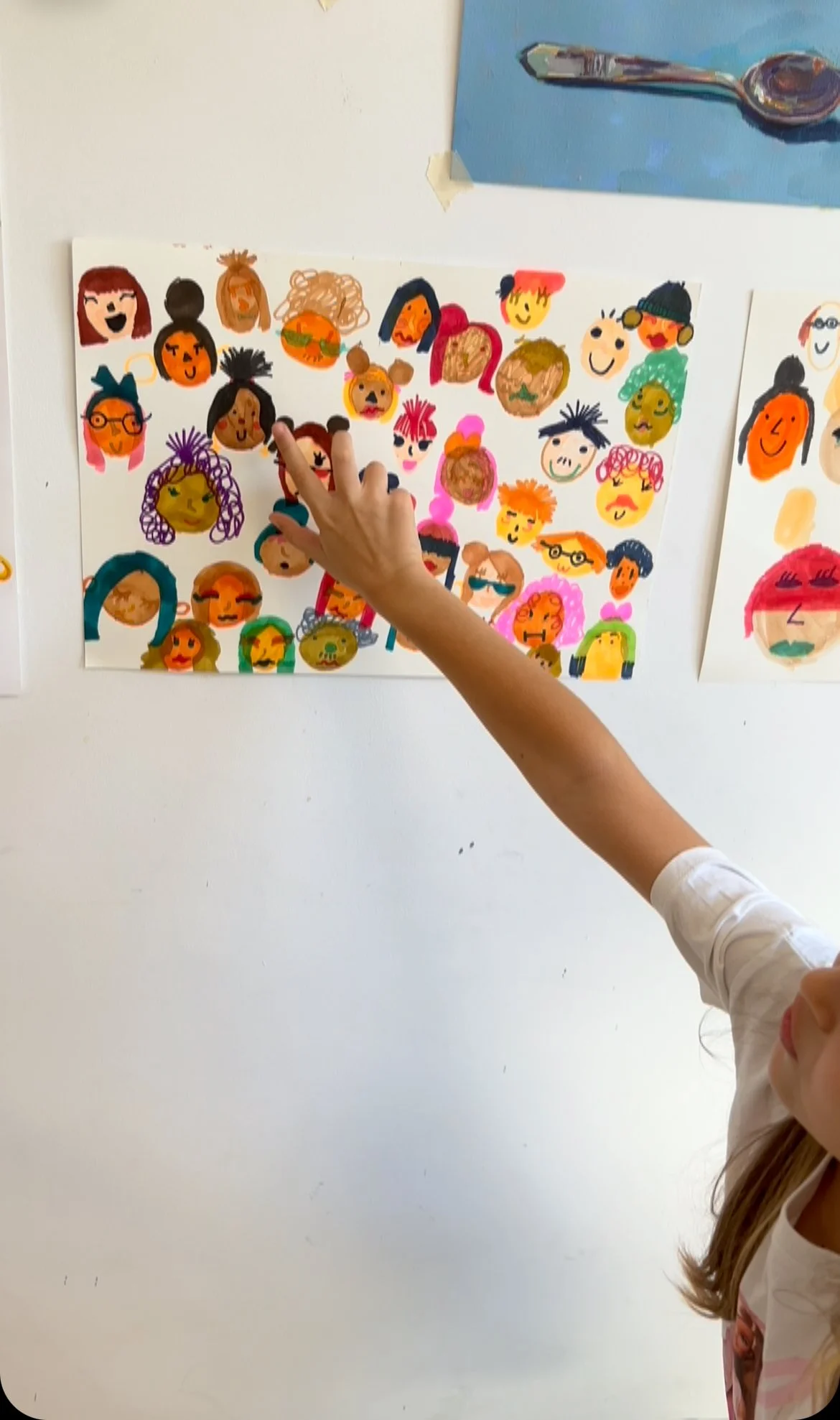 A child pointing at a colorful drawing of various animated-style faces on a white piece of paper taped to a wall.