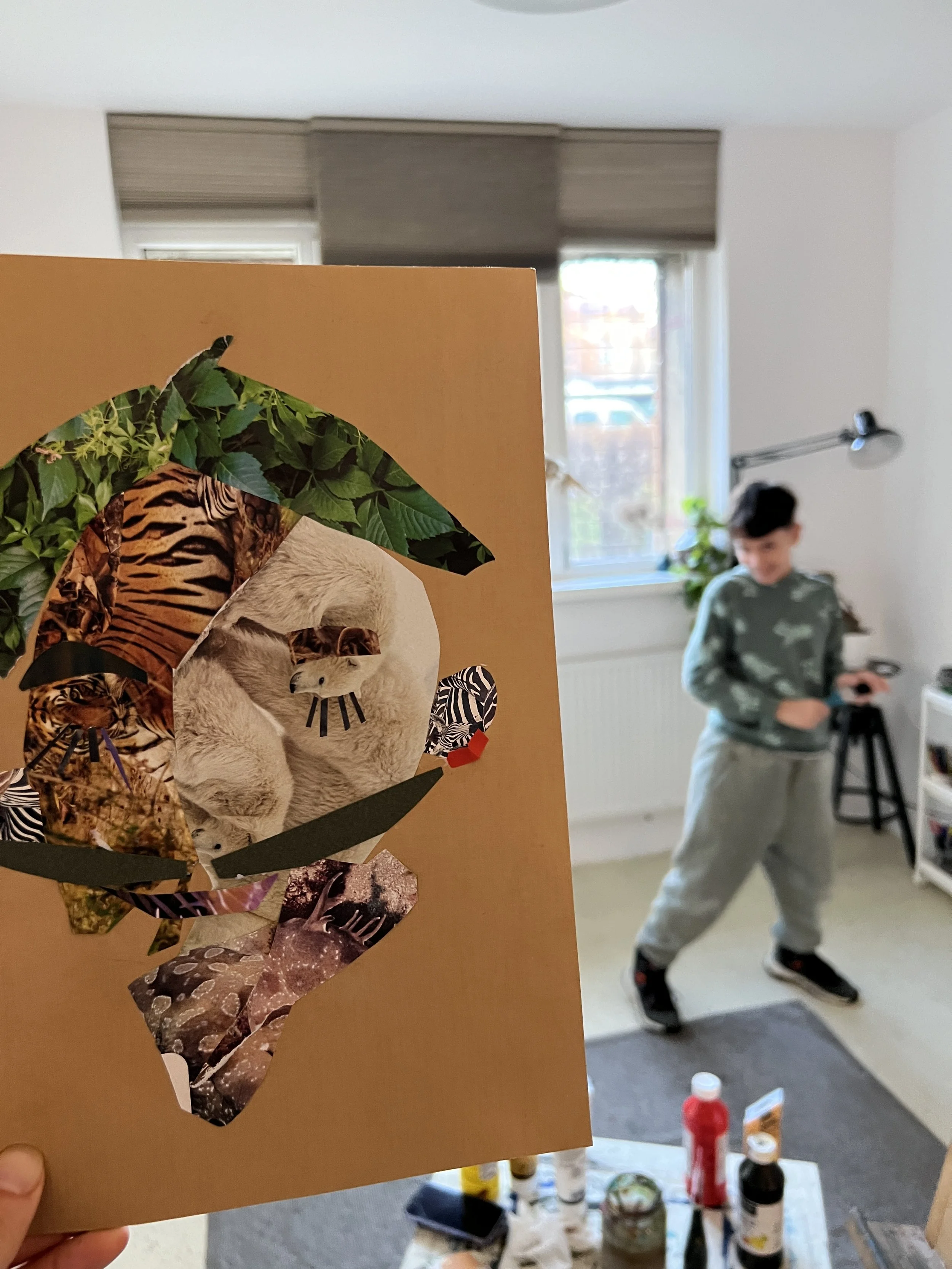 A collage artwork held close to the camera, featuring a central animal figure, possibly a dog, made from various patterned and textured paper cutouts, with surrounding nature-themed elements like leaves and animal prints.
