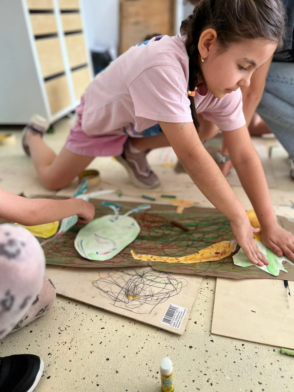 A young girl in pink shorts and a pink shirt is crawling on the floor, working on a large colorful drawing or craft project involving paper cutouts of animals, including a giraffe. She appears focused on her activity.