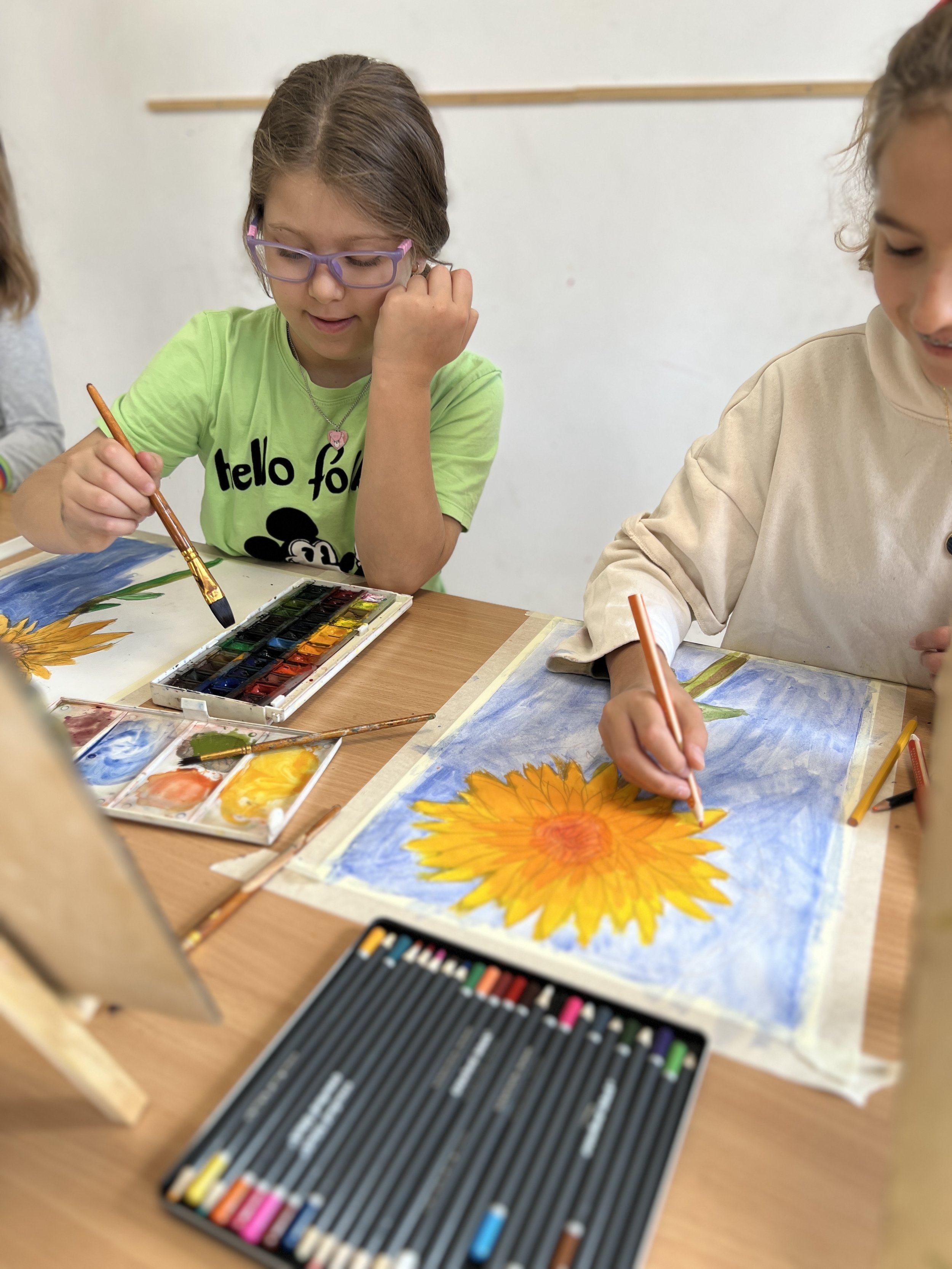 A girl wearing glasses and a green T-shirt is painting a sunflower with watercolors at a table, surrounded by painting supplies, in a classroom setting.