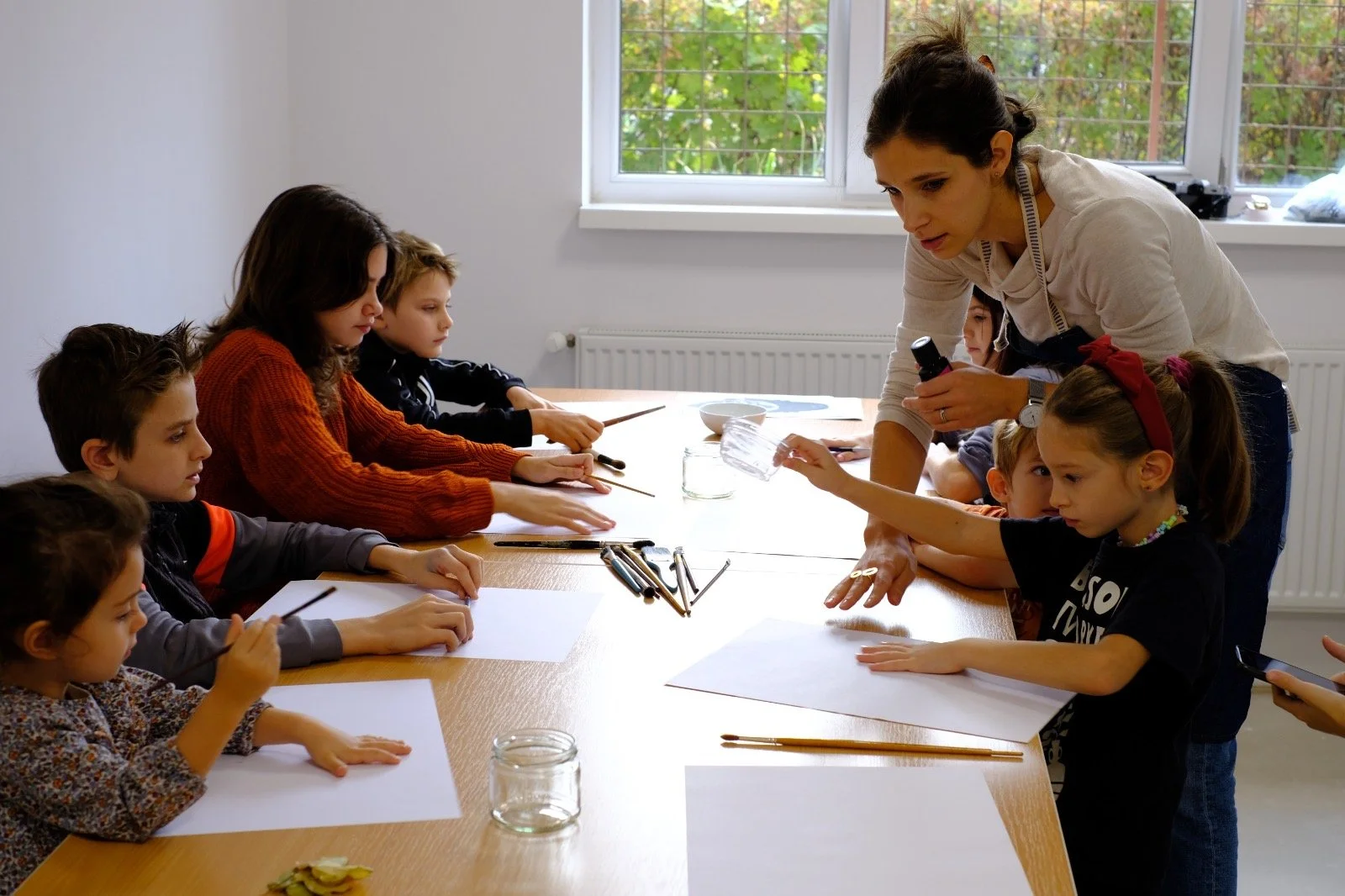 A female teacher showing art supplies to a group of children seated at a long table in a classroom, with papers and brushes on the table.