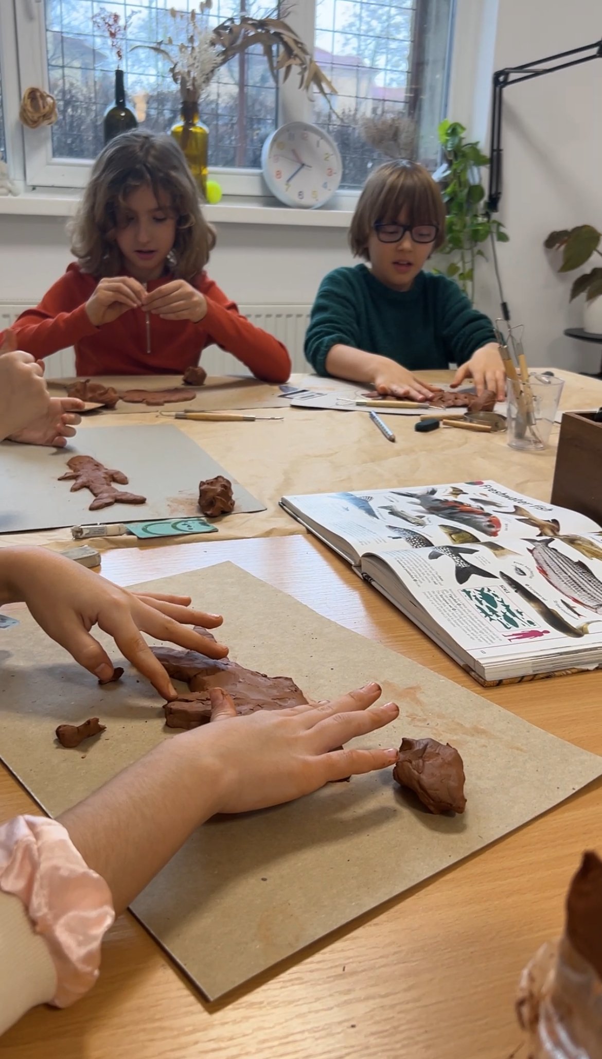 Children working with clay at a table during a craft activity, with an open book and craft tools on the table, and a window with decorative objects in the background.
